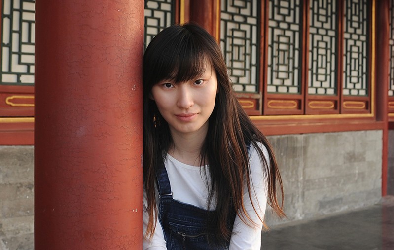 A girl sitting near a red, round stone pillar in front of a building.