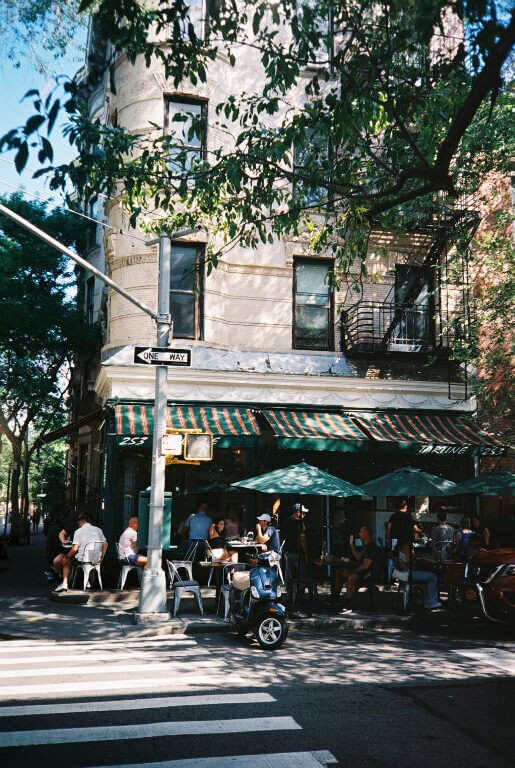 a film photo of a streetcorner in new york city with a cafe on the corner and people waiting on the sidewalk