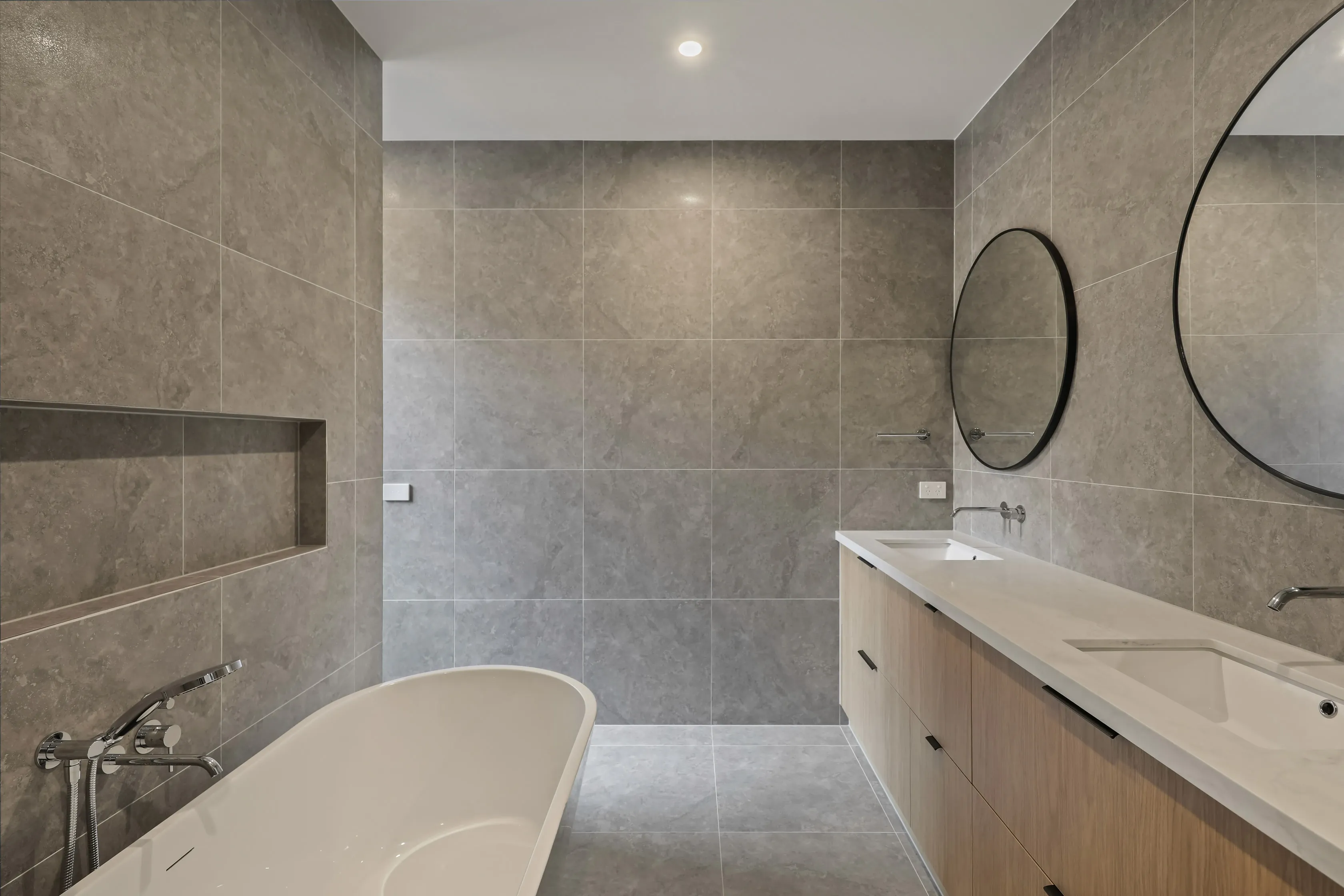 Ensuite bathroom at Mount Waverley Residence with grey stone tiles, freestanding bath, twin oak vanity and round black-framed mirrors