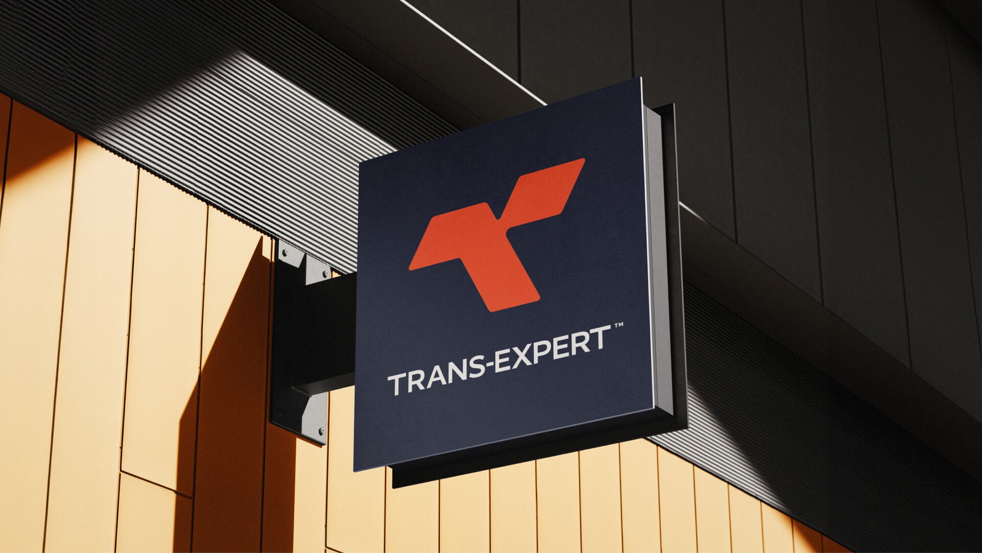 Modern outdoor wall sign with the Trans Expert logo in red and white on a dark blue background, mounted on a building with yellow and gray panels.
