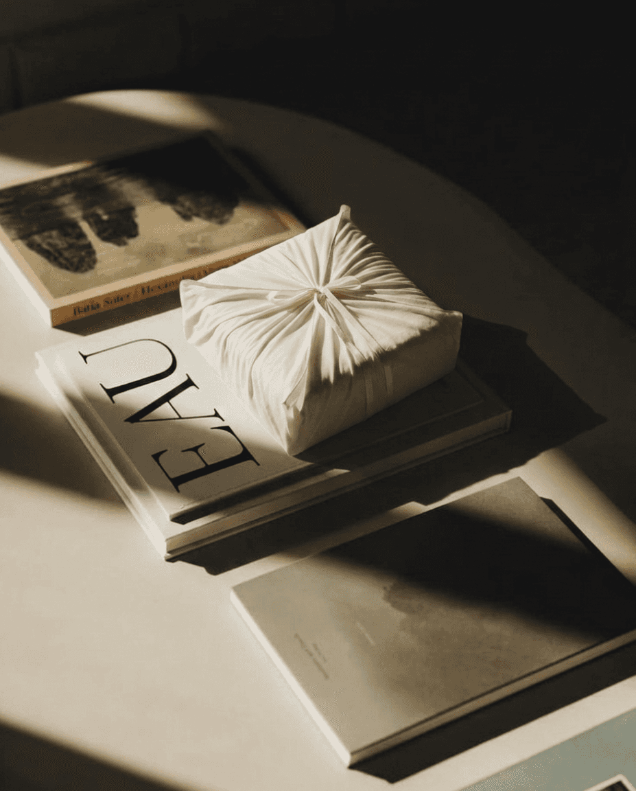 A neatly wrapped gift with white fabric sits on a stack of books titled "EAU" in sunlight. Warm tones and soft shadows create a serene, minimalist atmosphere.