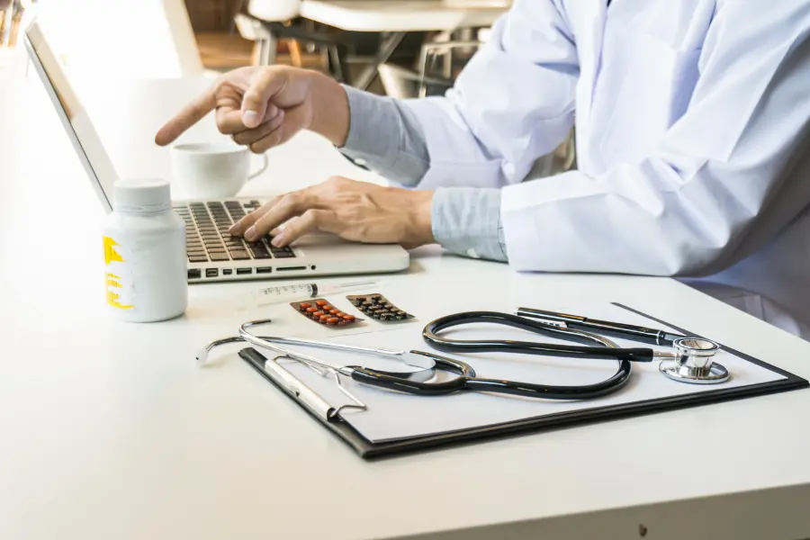 AI medical consulting captured as a white-coated doctor types on a laptop and points at the screen beside a stethoscope, pill bottle, and syringe on a clinic desk.