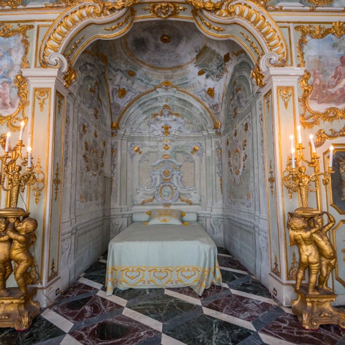 Luxurious Rococo-style room with ornate gold detailing, a canopied bed, marble floor, and golden candelabras held by cherub statues.