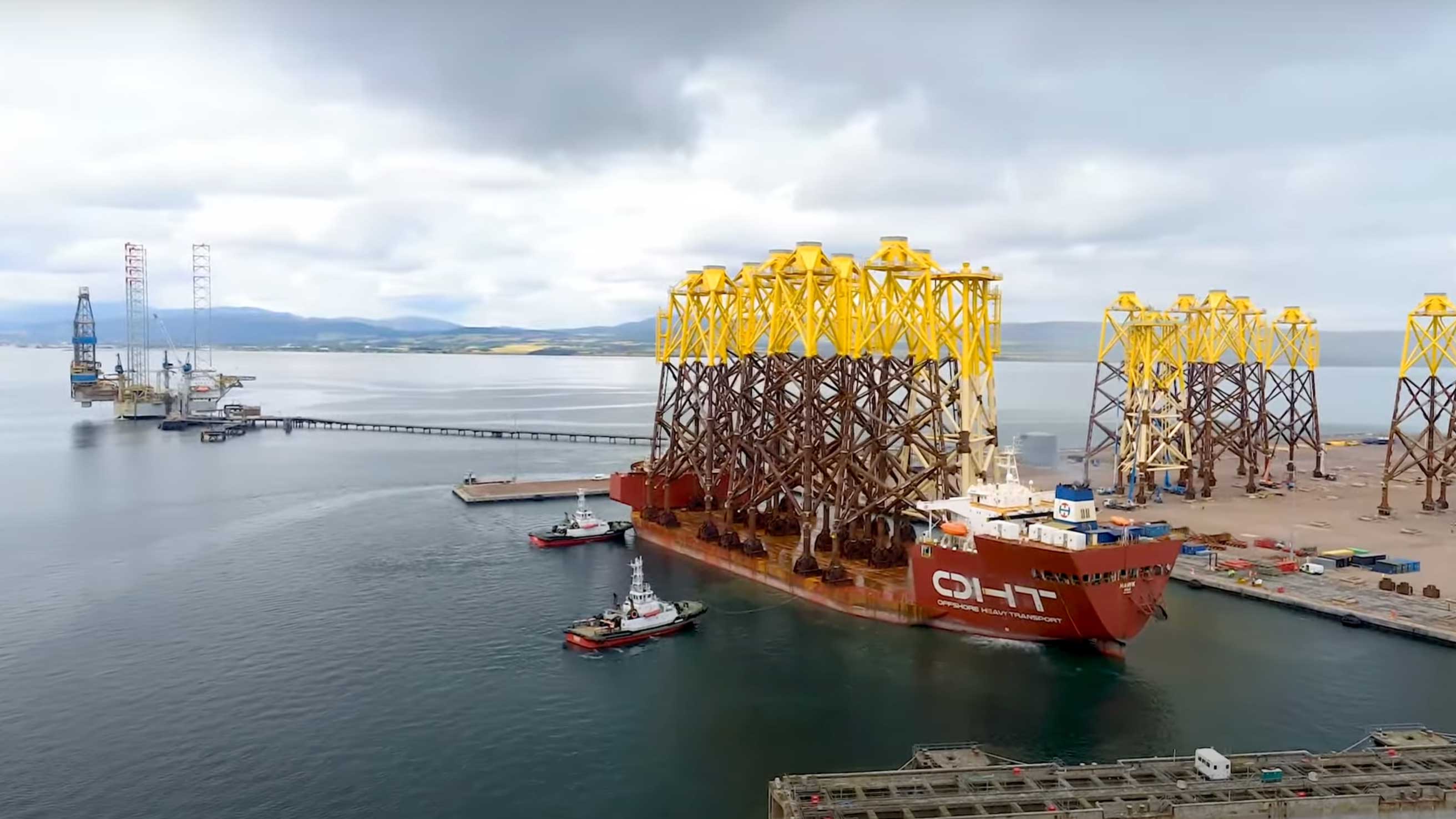 Offshore wind foundations loaded onto a vessel at a harbour with tugboats nearby.