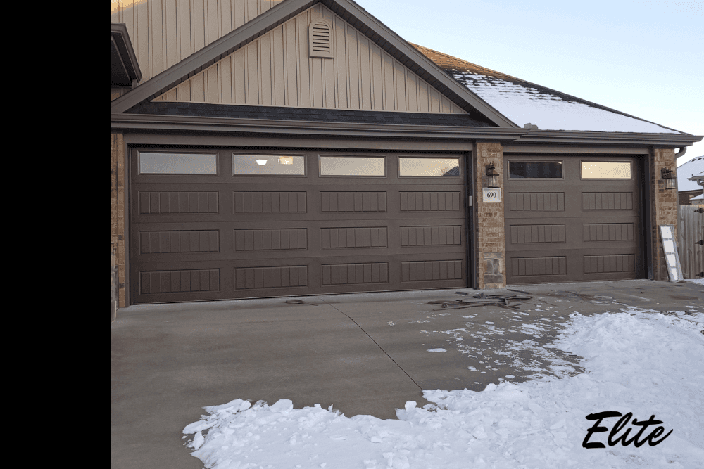 Brown double garage doors with snow along the driveway, showing winter exposure and potential cold weather wear.