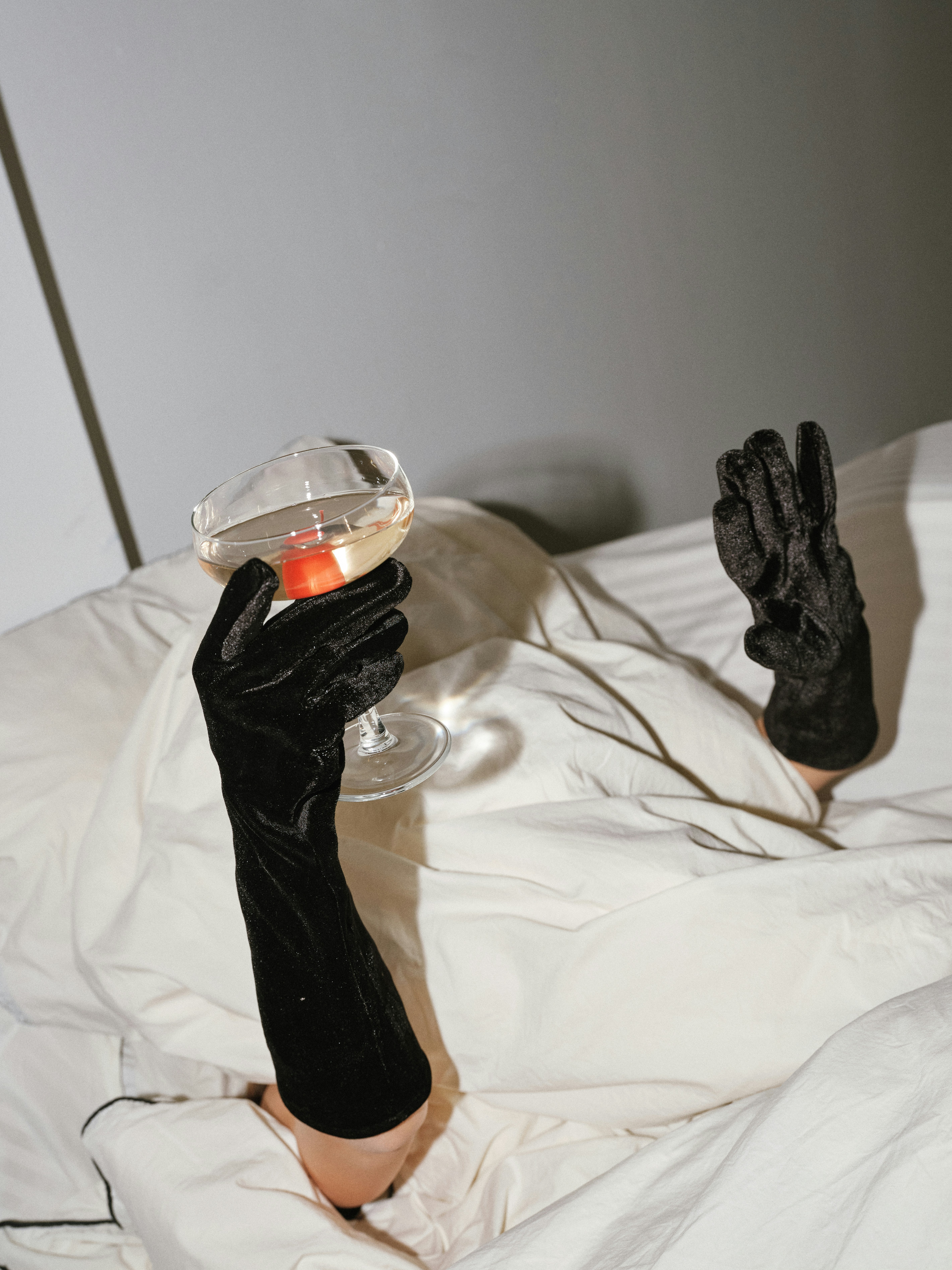 Conceptual editorial shot of two black velvet gloved arms emerging from white rumpled sheets, one hand holding a coupe glass with a red cherry, minimal grey background