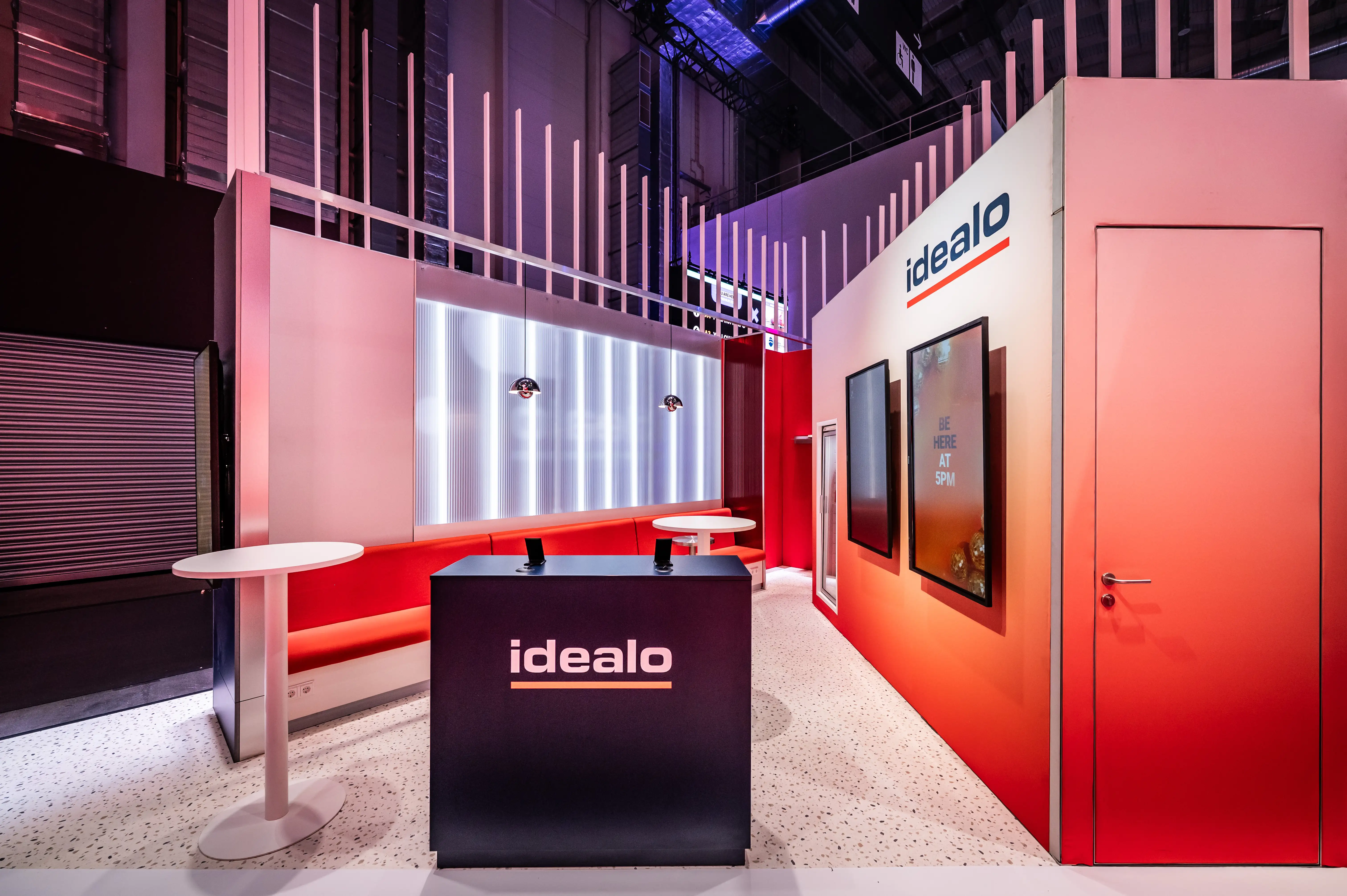 Idealo area with black counter, red gradient walls, screens and white bar tables under thin vertical poles.