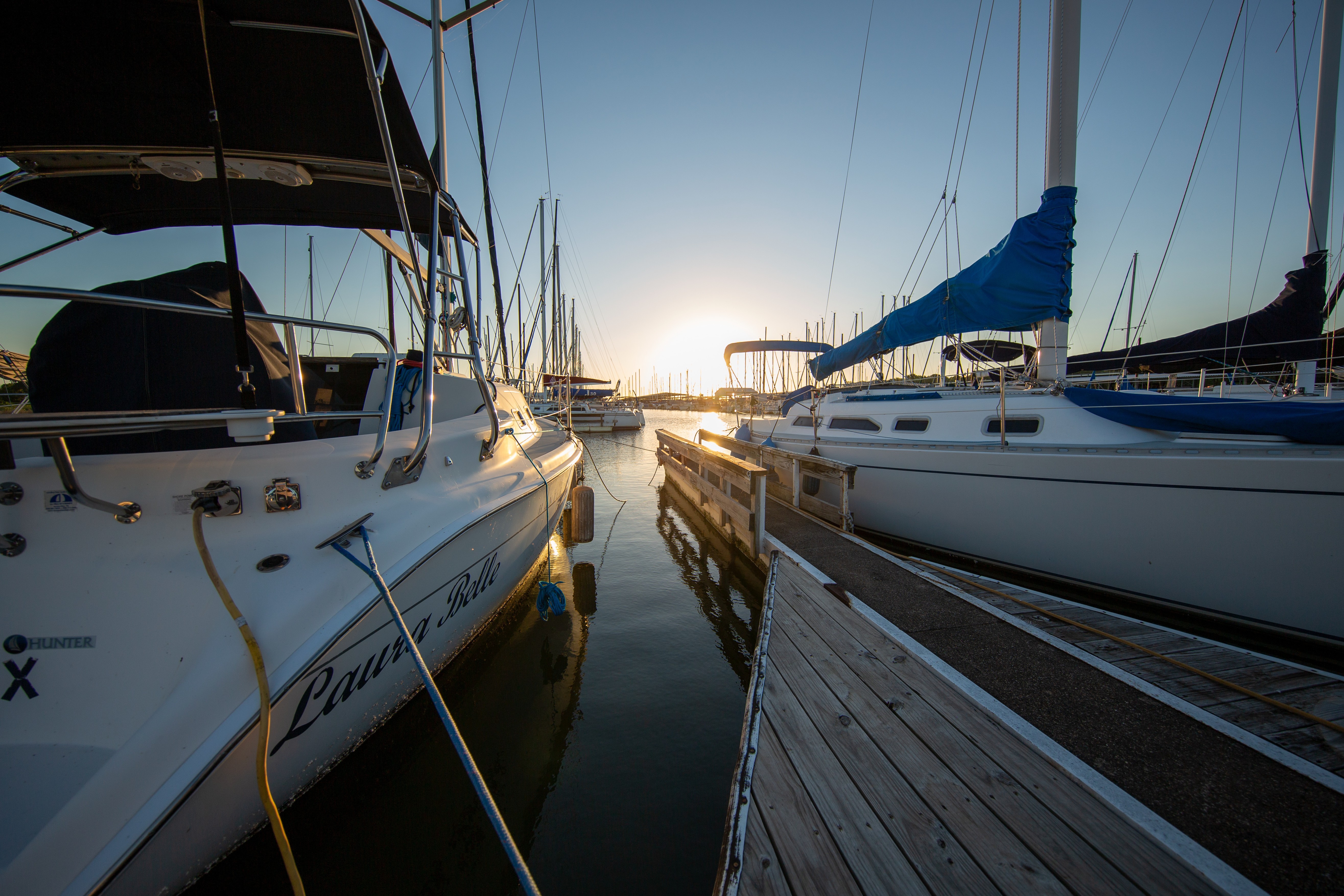 A serene marina scene at sunset featuring sleek white sailboats docked on calm waters, with a wooden pier leading into the horizon and masts silhouetted against the glowing sky.
