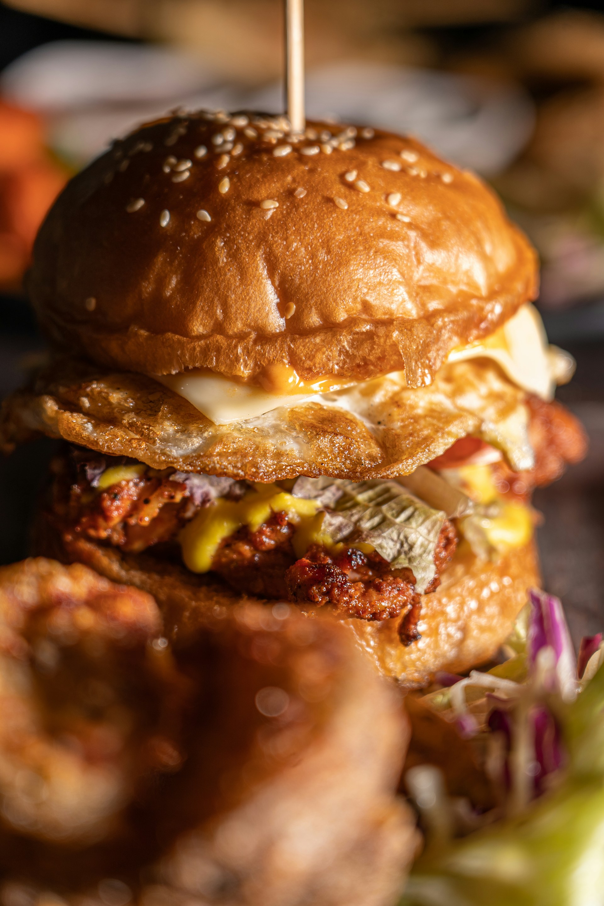 Close-up of a thick, appetizing burger layered with a fried egg, meat, cheese, and sauce, secured with a skewer.