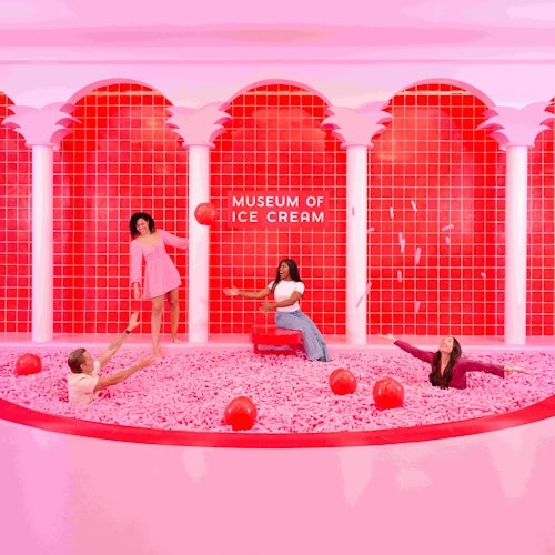 四个人在一个粉色球池中玩乐和摆姿势，球池里有红色的球，拱门上写着“冰淇淋博物馆”。