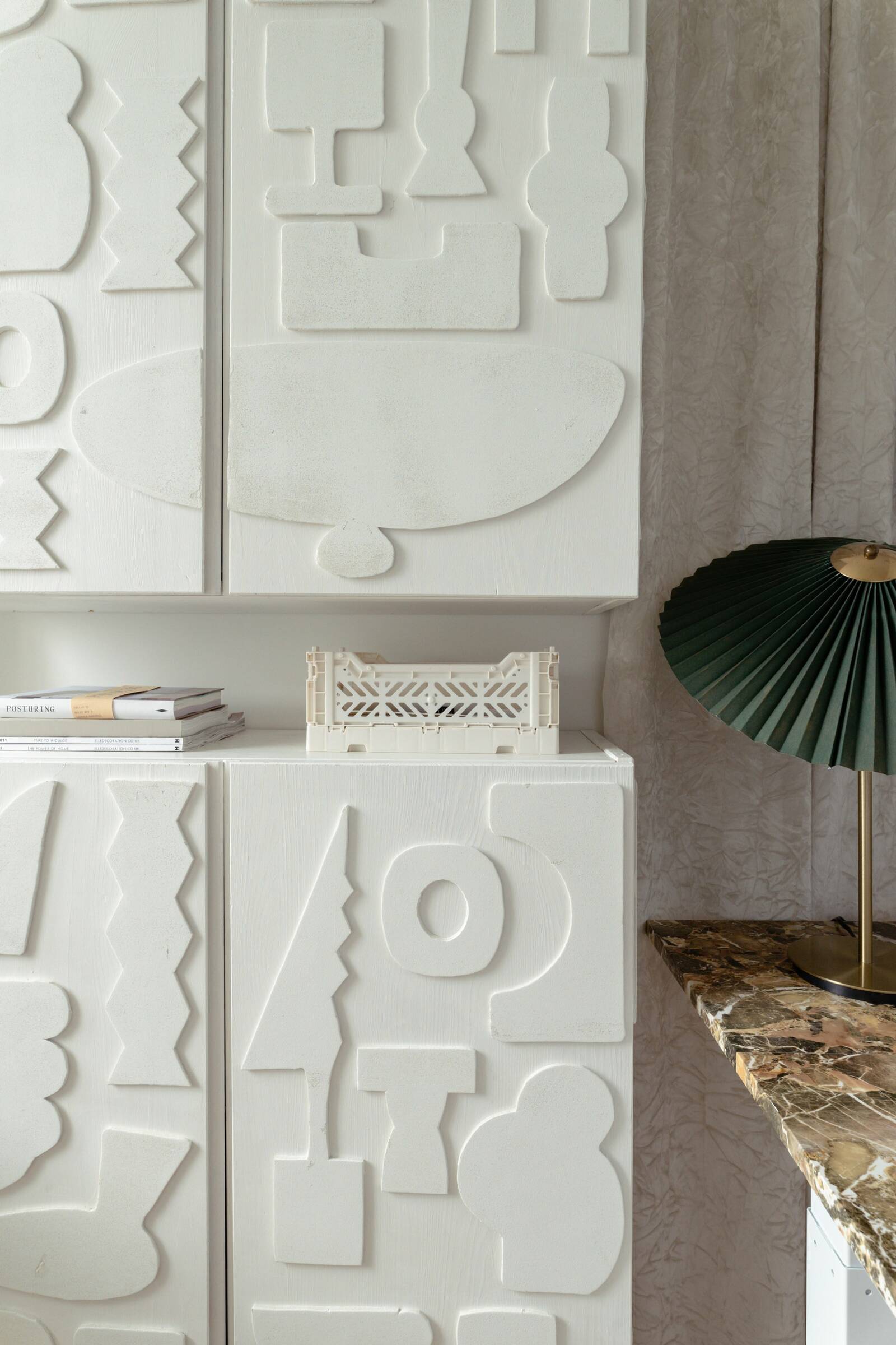 Close-up of a white textured storage with abstract geometric shapes, a green lighting fixture on a marble table, and a small stack of books.