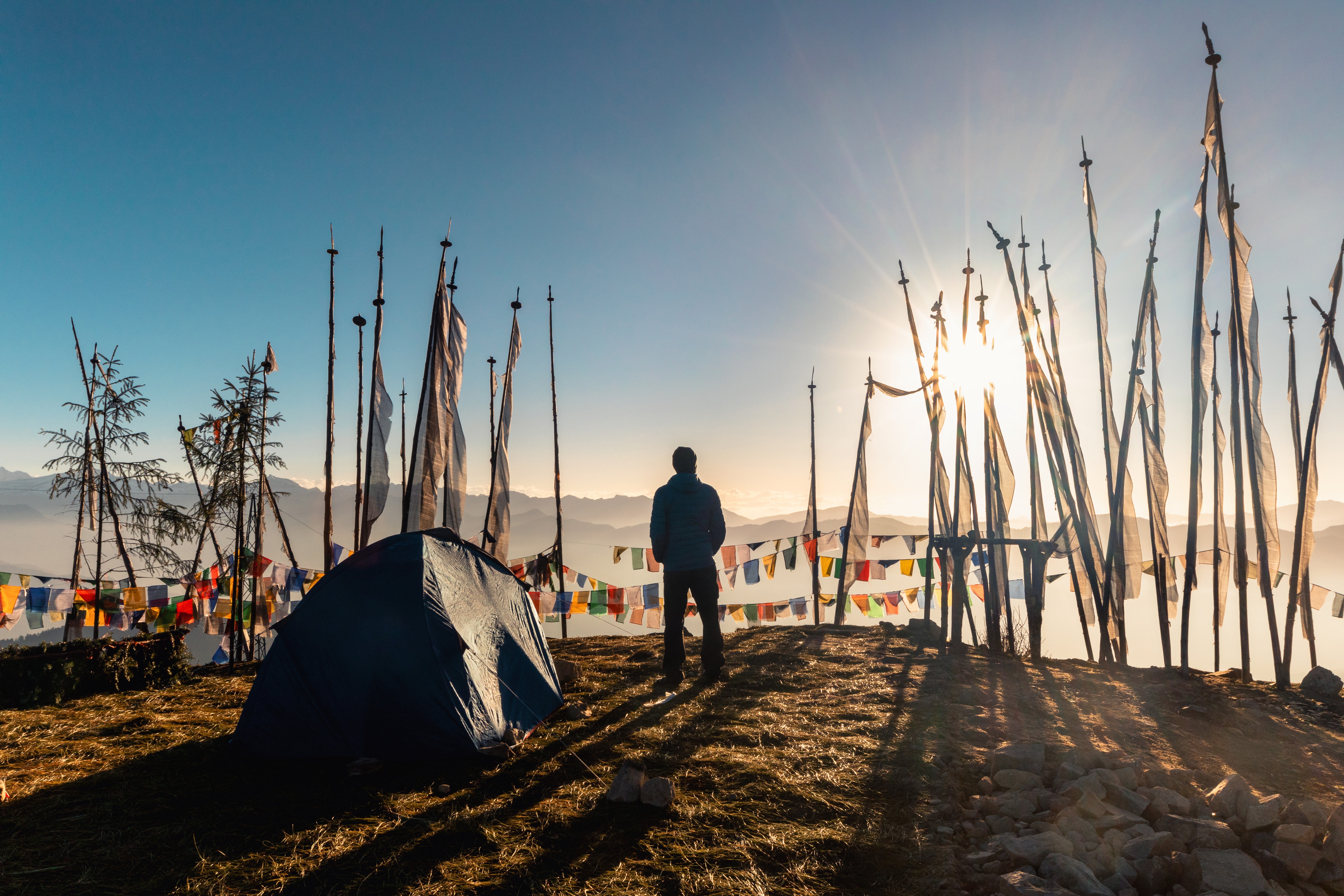 A camper stands beside a tent on a mountain ridge at sunrise, surrounded by tall prayer flags and colorful prayer banners fluttering in the light.