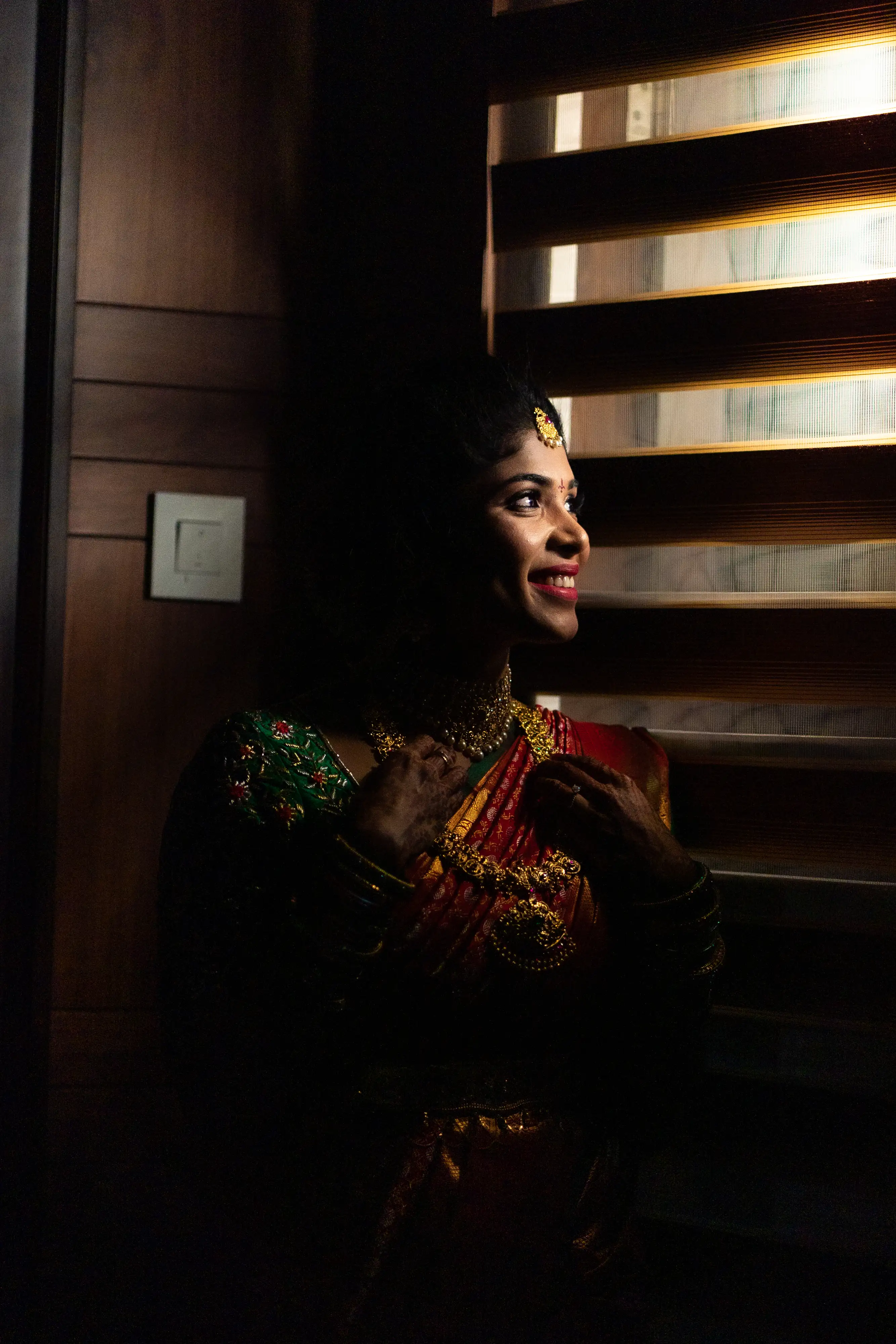 Harshitha, the bride, gazes through window blinds in an artistic pose. Photo by Out of The Blues Fine Art Wedding Photography in Hyderabad.