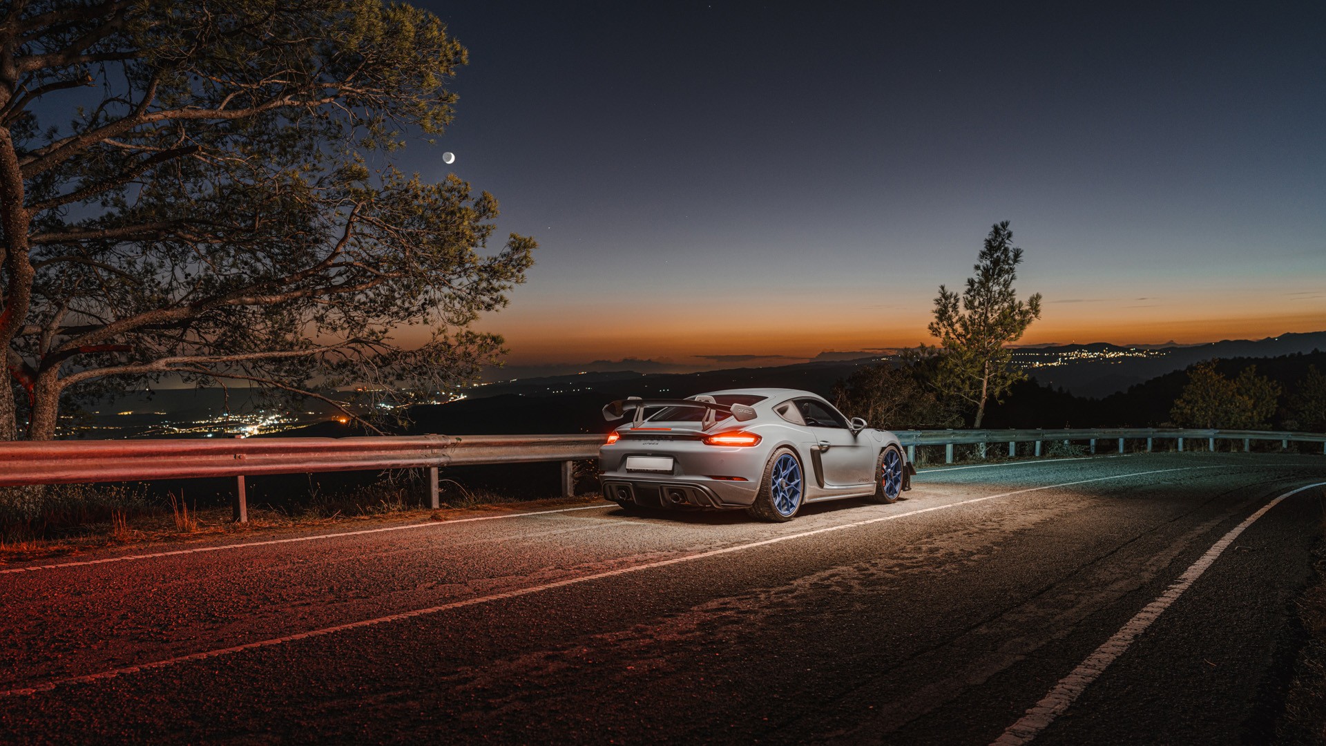 Grey Porsche 718 Cayman GT4 RS with blue wheels seen from the rear three-quarter, parked at the roadside on a mountain pass at twilight, rear wing and brake lights in profile, with a crescent moon and a glowing horizon over the hills of Cyprus.