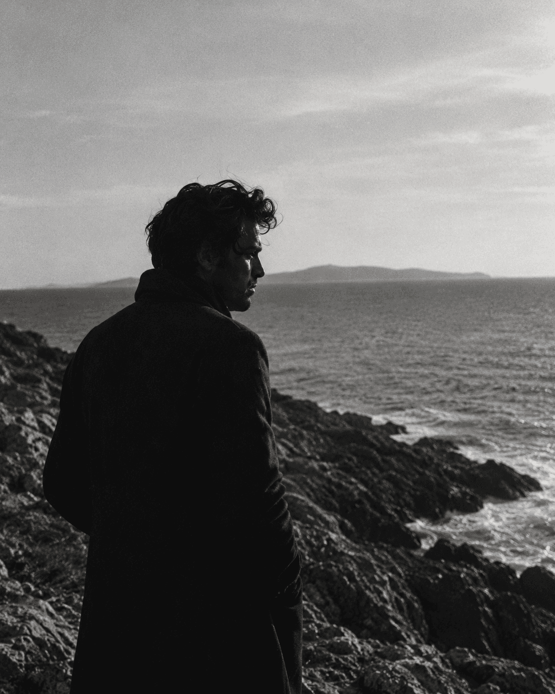 Black-and-white photograph of a man looking out over the sea from a rocky coastline.