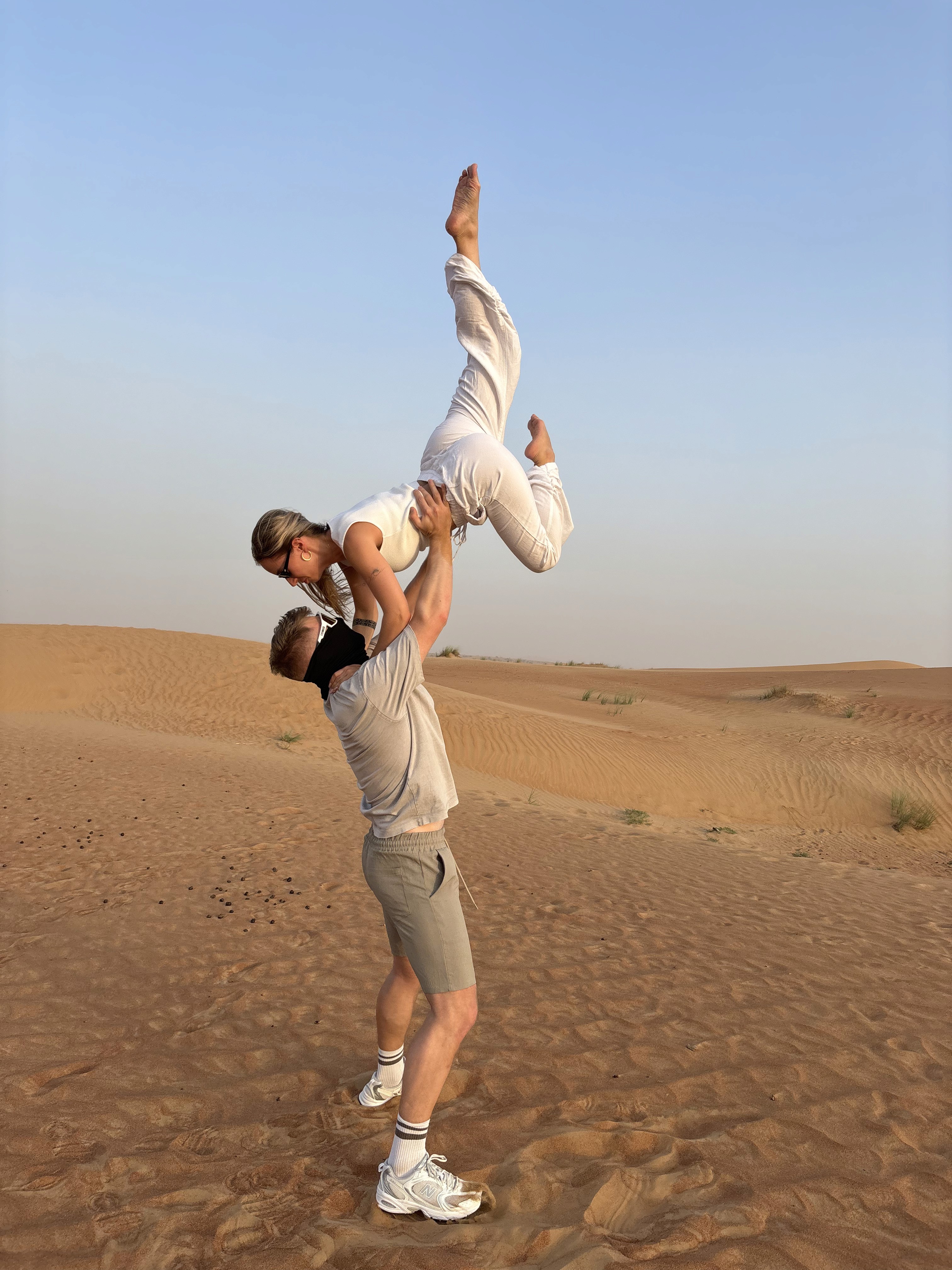 A woman with long blonde hair, in a brown top and jeans, walks through a sunlit forest, holding someone's hand. The scene is warm and inviting.