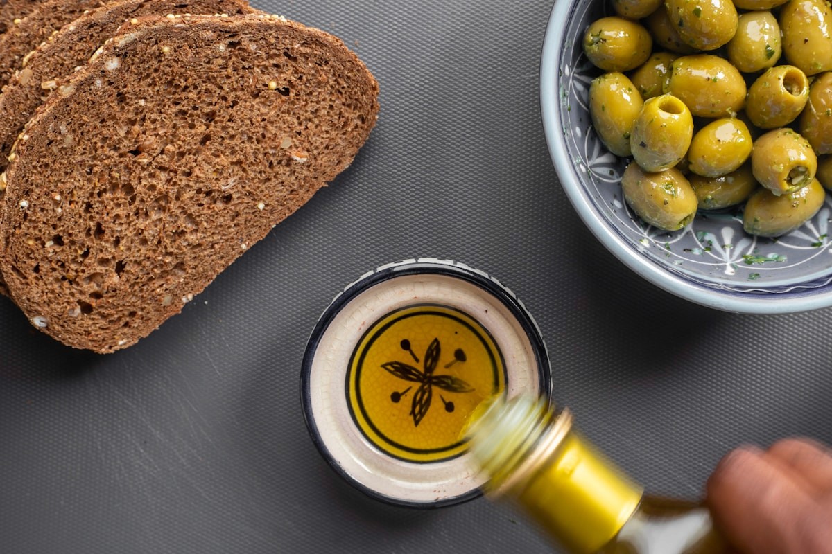 Slices of whole grain bread, a bowl of green olives, and olive oil being poured, staples of a Mediterranean-style anti-inflammatory diet