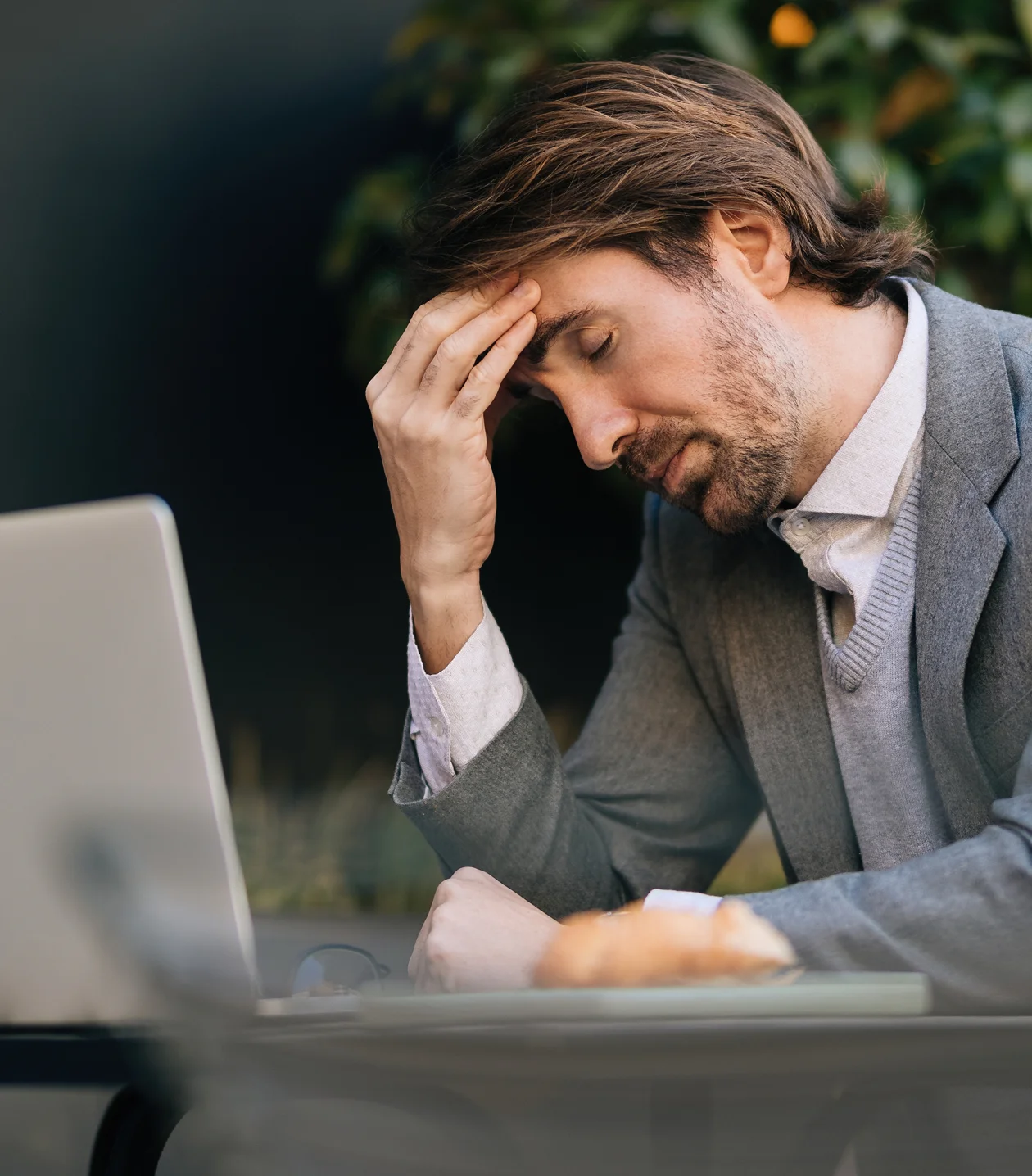 Stressed man in a grey blazer sits at a laptop with his eyes closed and hand on his forehead.