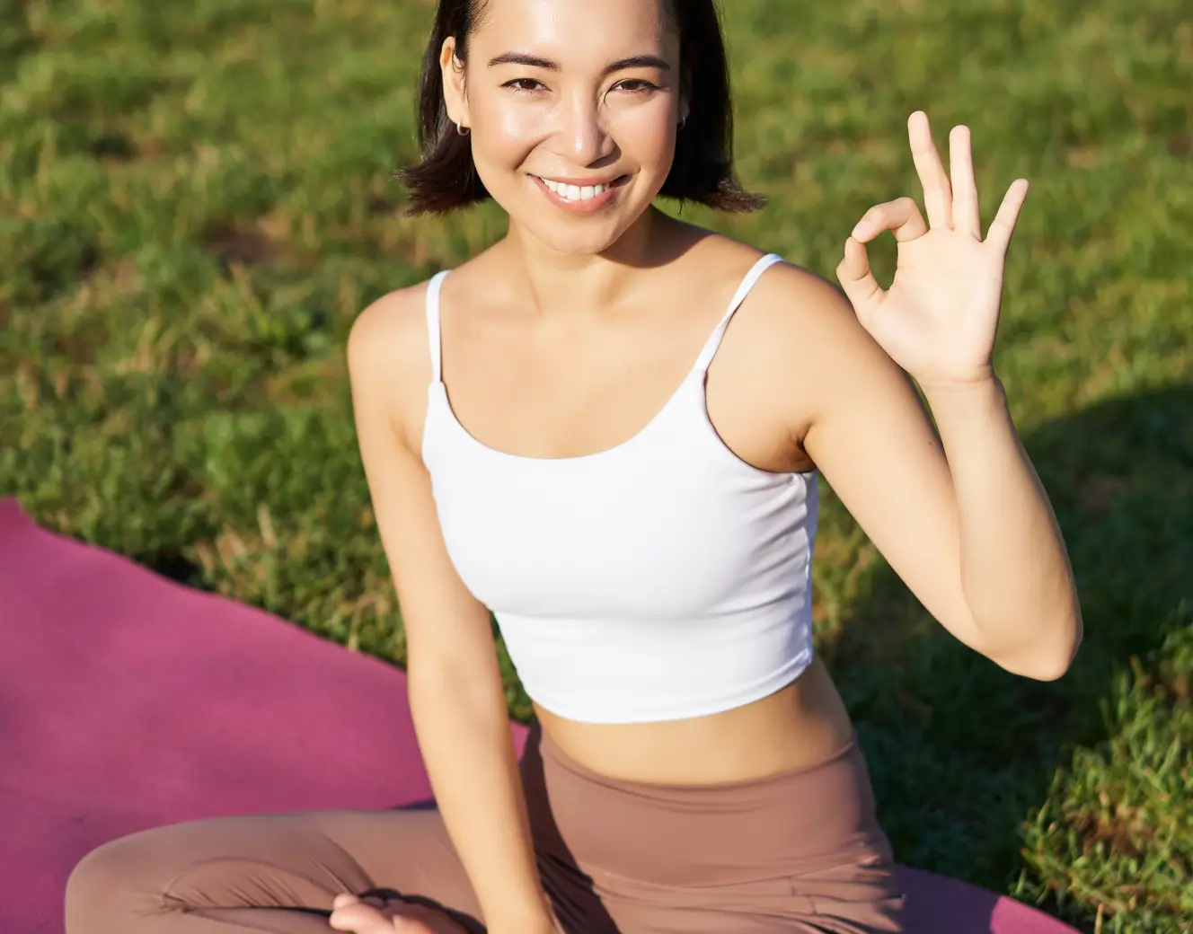 Smiling woman sitting cross-legged on a yoga mat outdoors showing an OK hand gesture.