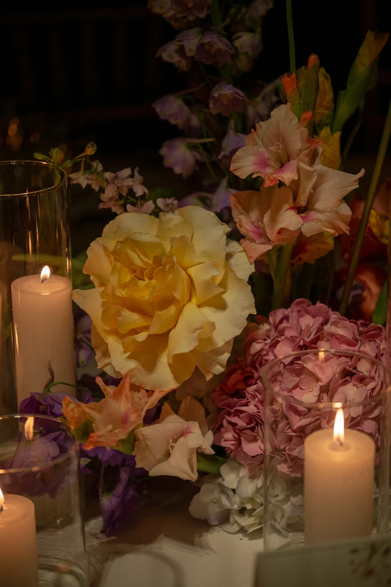Primer plano de rosas amarillas, hortensias rosas y delphiniums iluminados por velas en mesa de recepción.