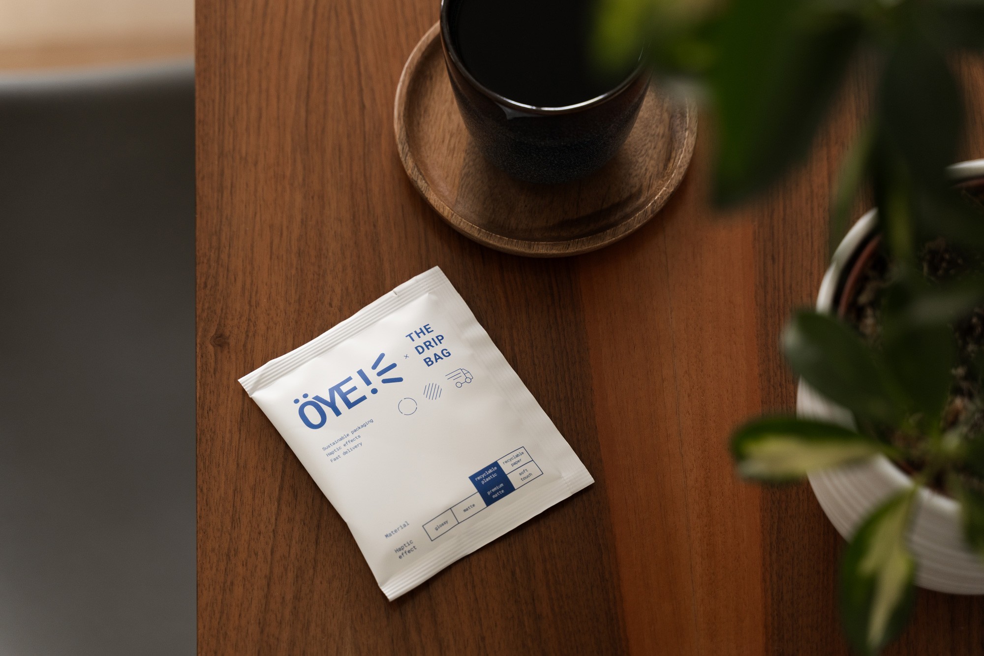 OYE! drip kafijas maisiņš uz galda ar krūzi — minimālistiska dzīvesstila produktu fotogrāfija, kas uzsver ilgtspējīgu iepakojumu un mājīgu atmosfēru.