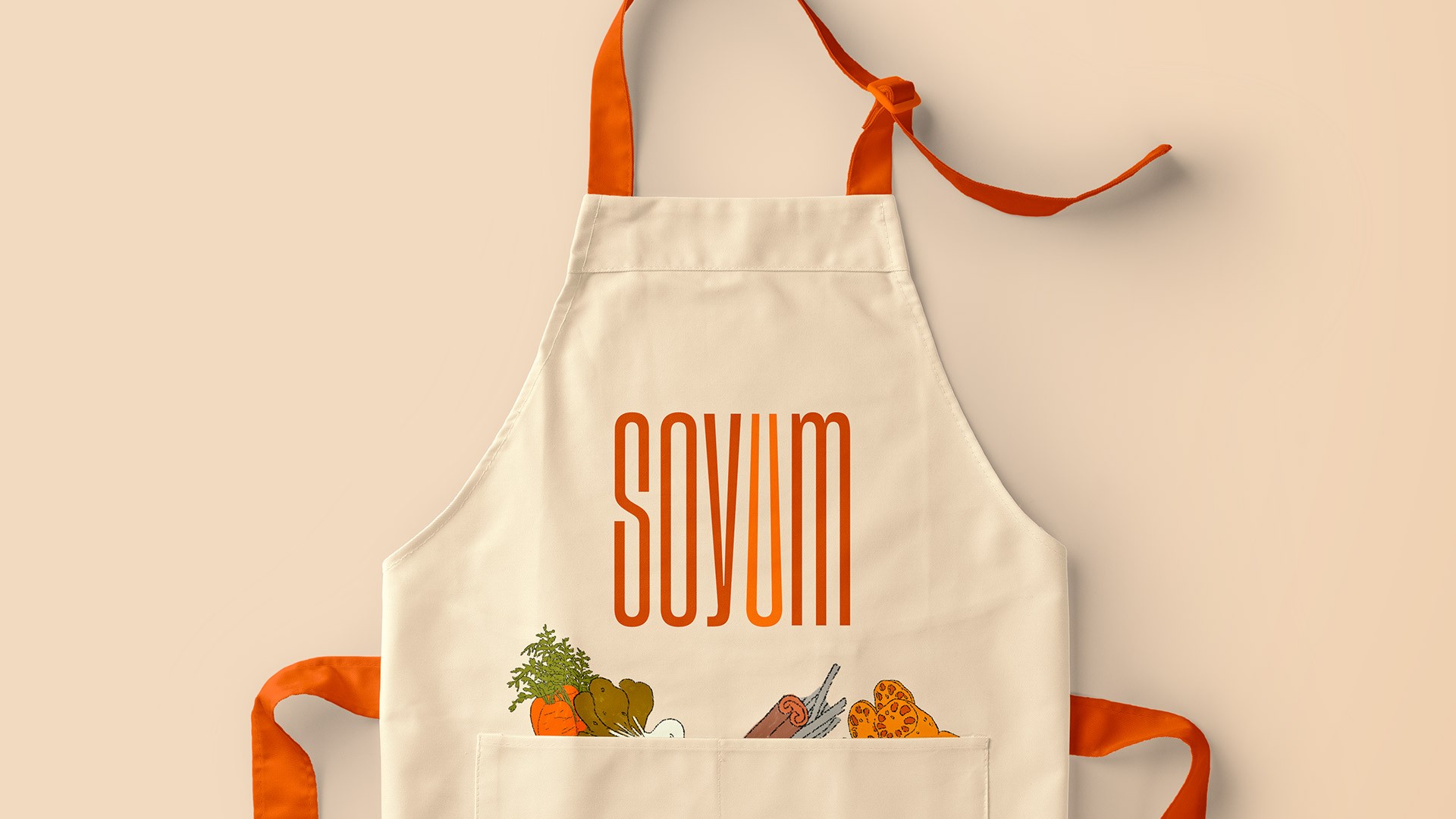 A white apron with orange straps and ties features the word "SOVUM" in bold orange letters across the chest. Below the lettering, a small pocket on the apron holds a collection of colorful vegetables, including carrots, broccoli, and leafy greens. The apron is presented against a light peach-colored background.