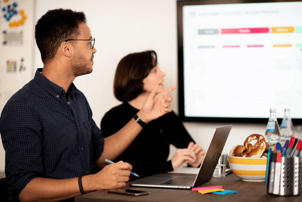 Mann und Frau sitzen in einem Meetingraum und schauen auf einen großen Bildschirm mit einer Prozessübersicht. Der Mann gestikuliert und spricht, während die Frau zuhört. 