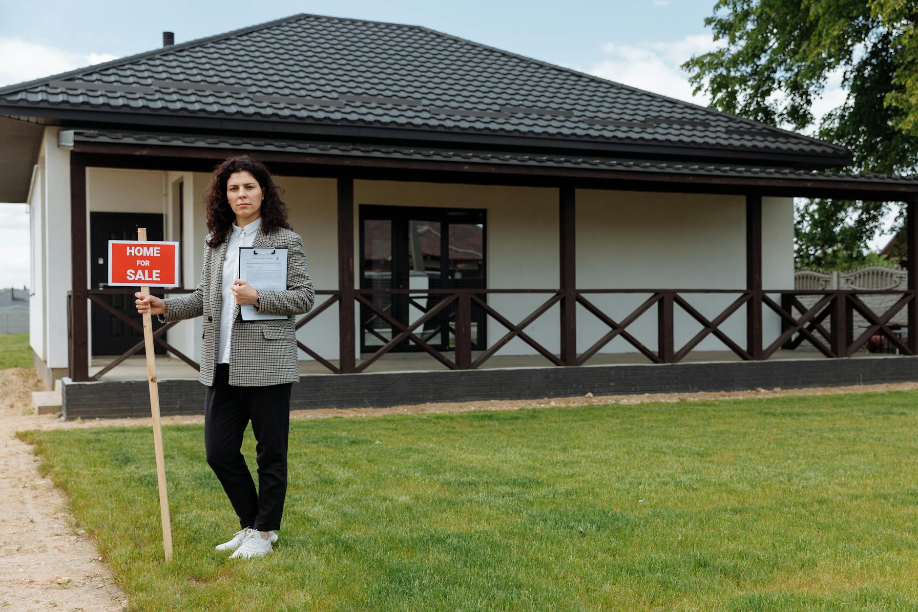 Woman in grey blazer holding a clipboard and Home For Sale sign on a lawn in front of a white bungalow.