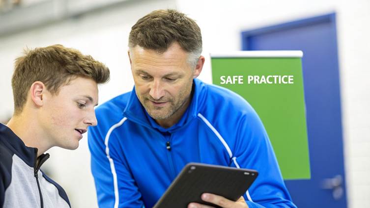 Two men, possibly a coach and athlete, look at a tablet with a 'SAFE PRACTICE' sign behind them.