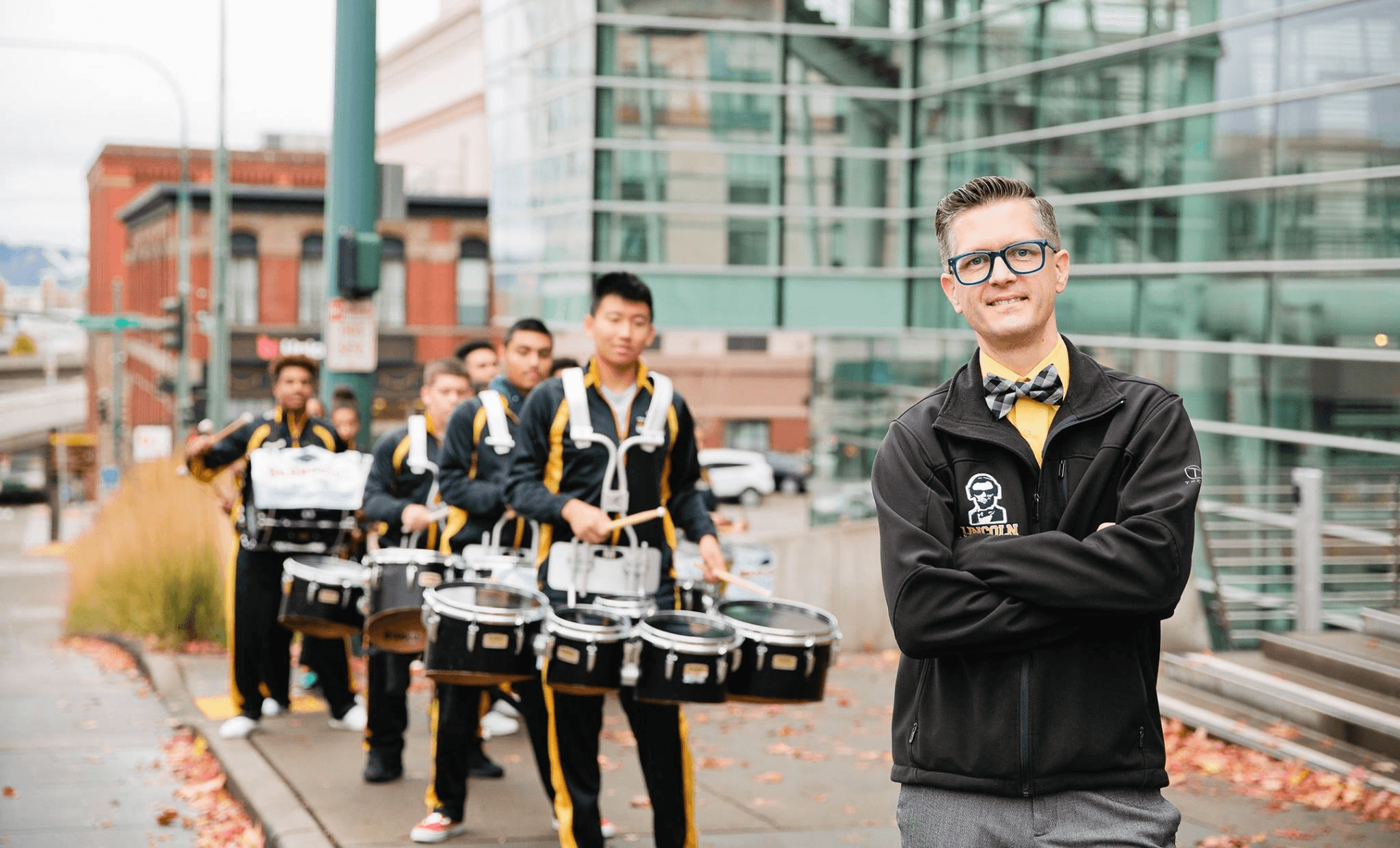 Peter Briggs stands on Broadway in Tacoma in front of his Lincoln High School drumline — arms crossed, smiling — students in black and gold uniforms playing drums behind him