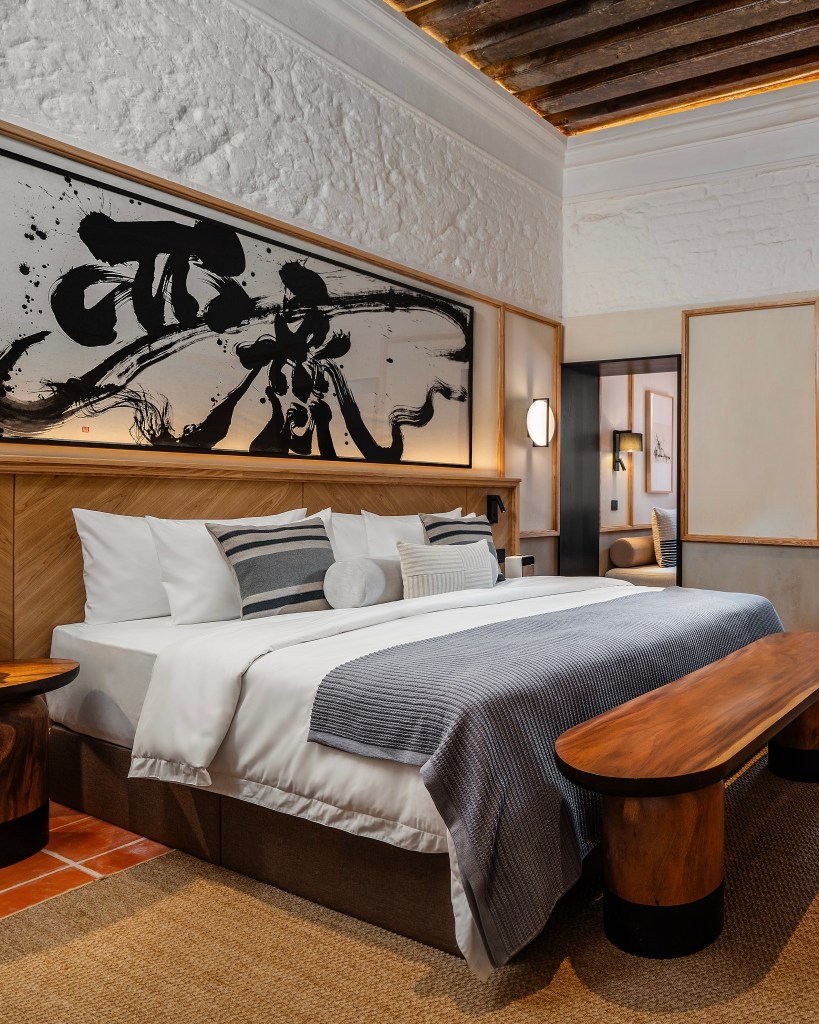 A stylish hotel room featuring a king-size bed with decorative pillows and a cozy blanket, complemented by wooden furniture and an artistic wall hanging.