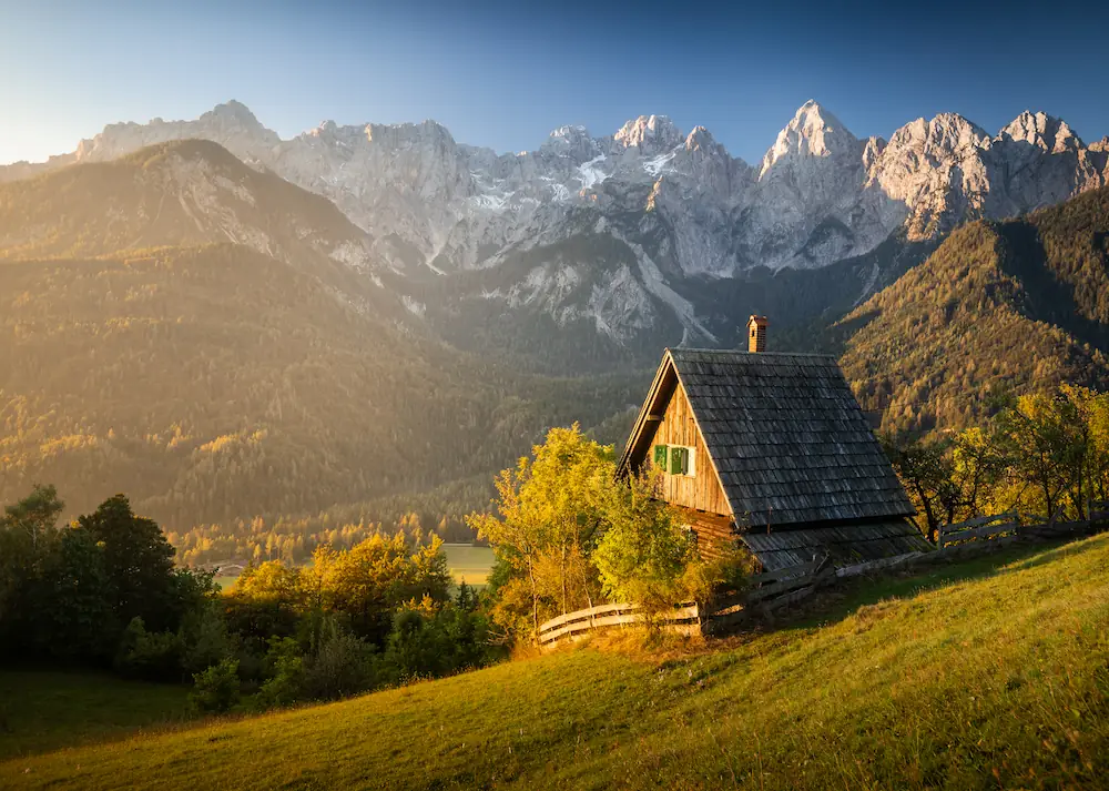 A rustic mountain hut in Srednji Vrh, Slovenia, overlooking a deep valley and the dramatic, sun-drenched peaks of the Julian Alps.