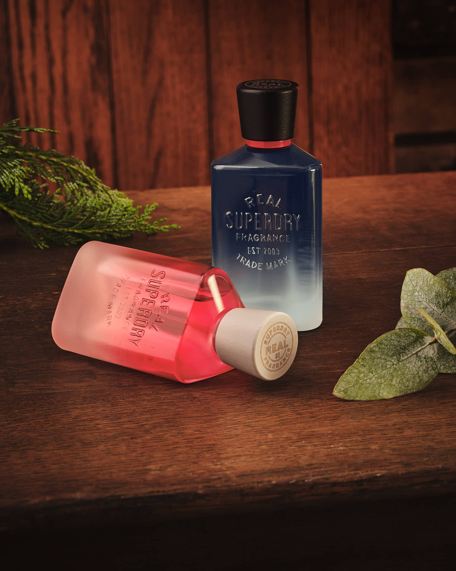 Two bottles of perfume, one red and one blue, displayed on a wooden surface with green leaves.