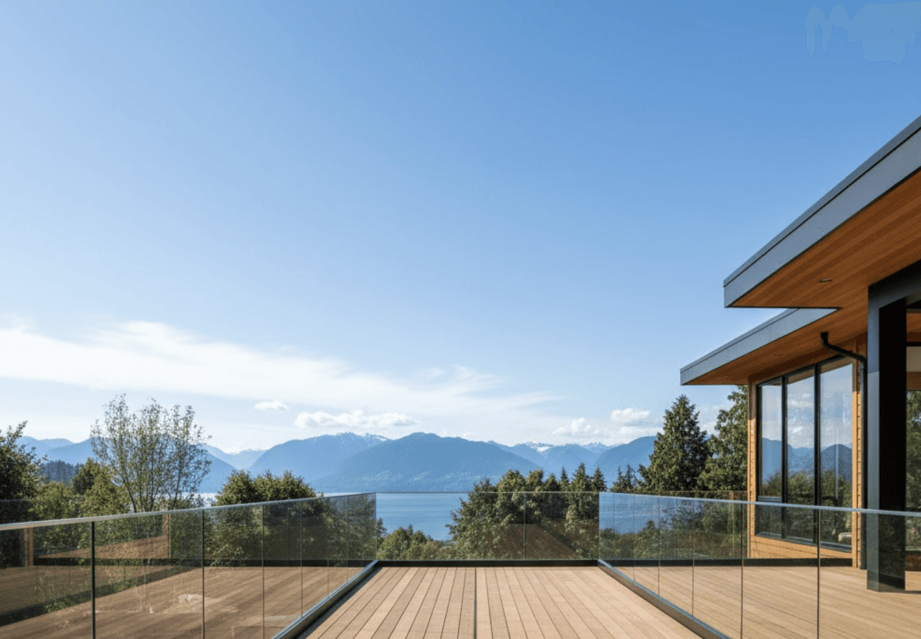 A modern Vancouver home with frameless glass railings on a sunny day — sleek architecture overlooking mountains and ocean. The glass railings wrap around a spacious wooden deck with clear reflections of the sky. The scene captures natural light streaming through the transparent panels, showing how the design preserves unobstructed views and creates a bright, open atmosphere.