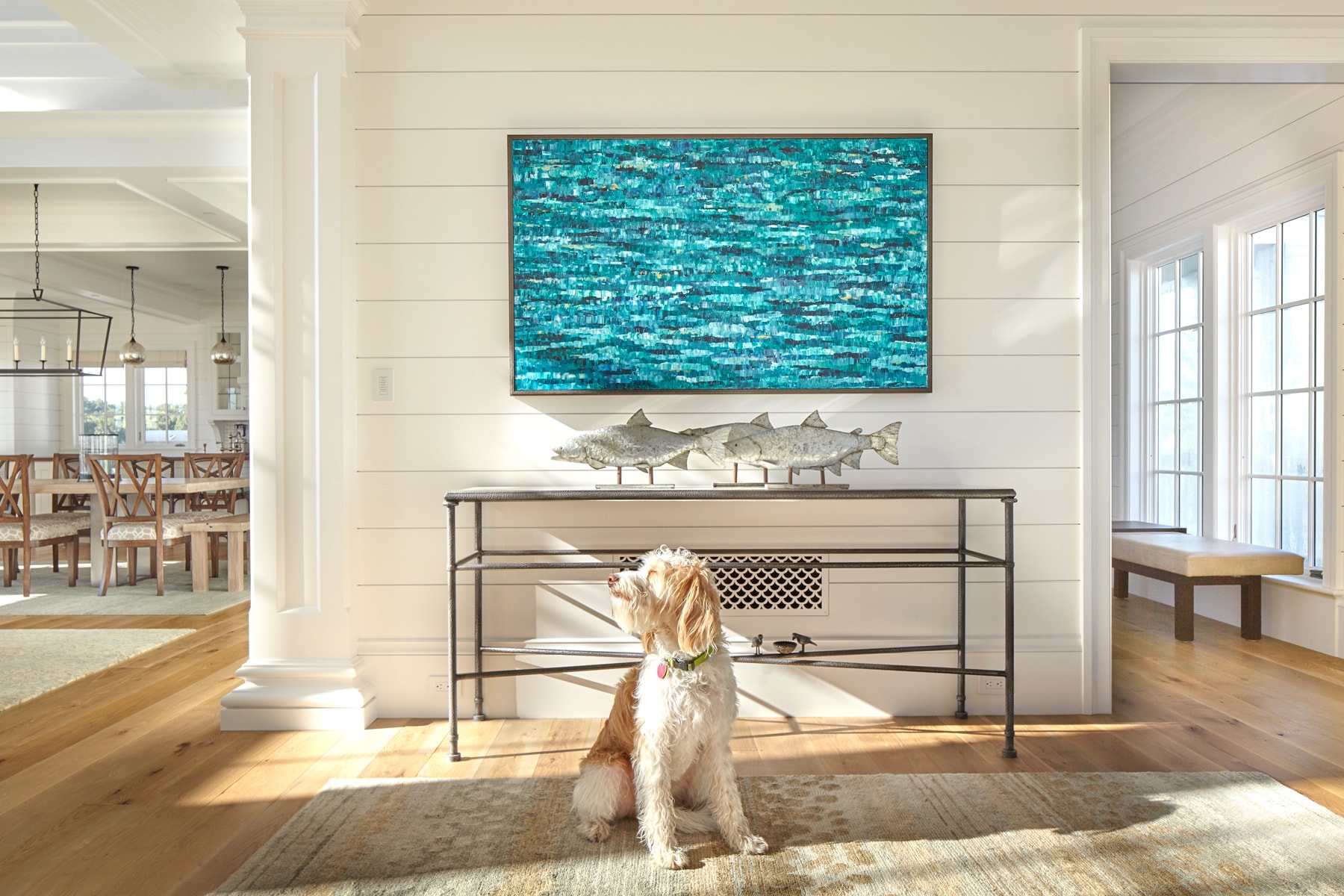 A white coastal-style foyer with a wooden floor, featuring a small dog, a console table with fish sculptures, and a large abstract turquoise painting.