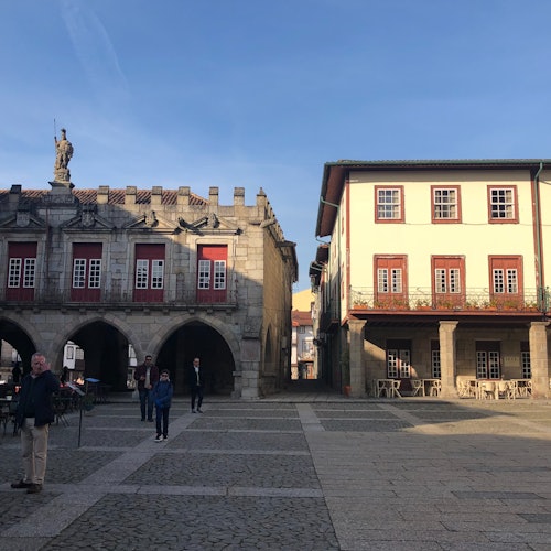 一個市鎮廣場，有一座歷史建築，左側有拱門，右側有一棟紅窗框的兩層樓建築。人不多。