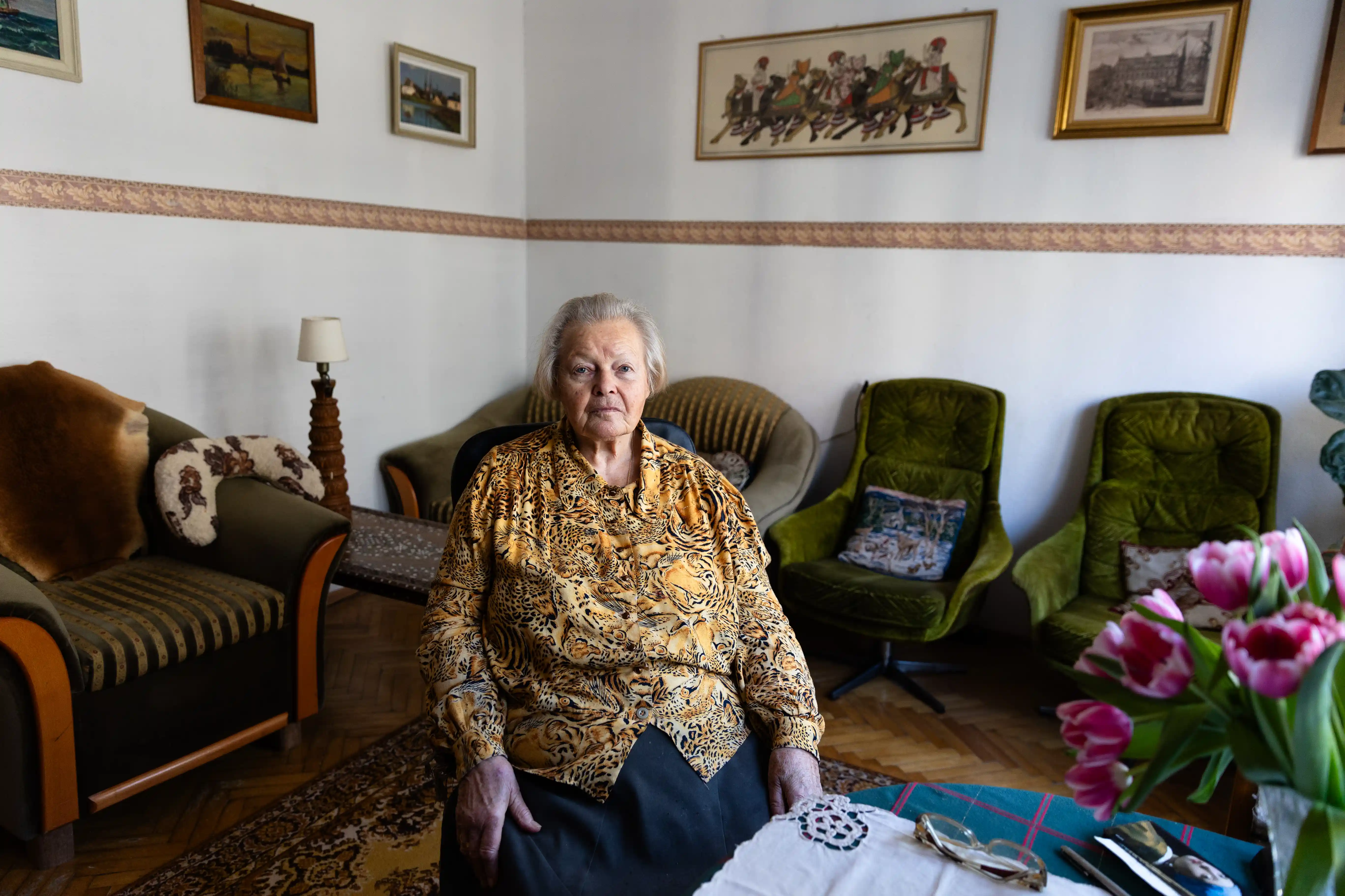Starsza kobieta w koszuli siedzi w fotelu. Za nią widnieje salon z wieloma fotelami oraz obrazami na ścianach.