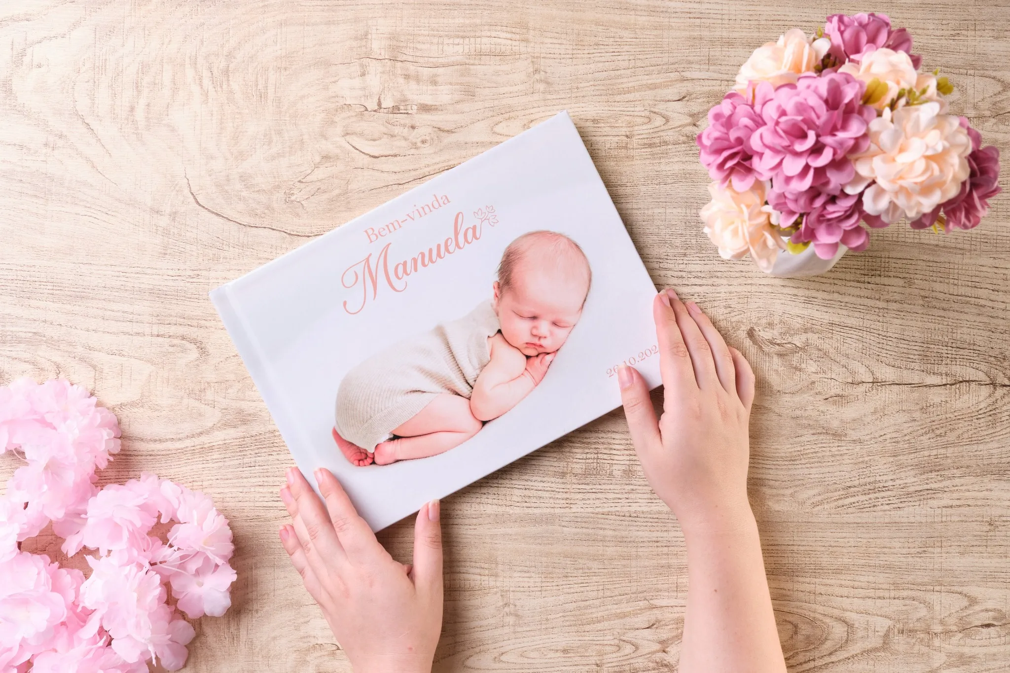 Mãos segurando fotolivro com capa personalizada de duas mulheres sorrindo, com a frase “Eu amo o Nordeste”, sobre mesa de madeira e flores decorativas