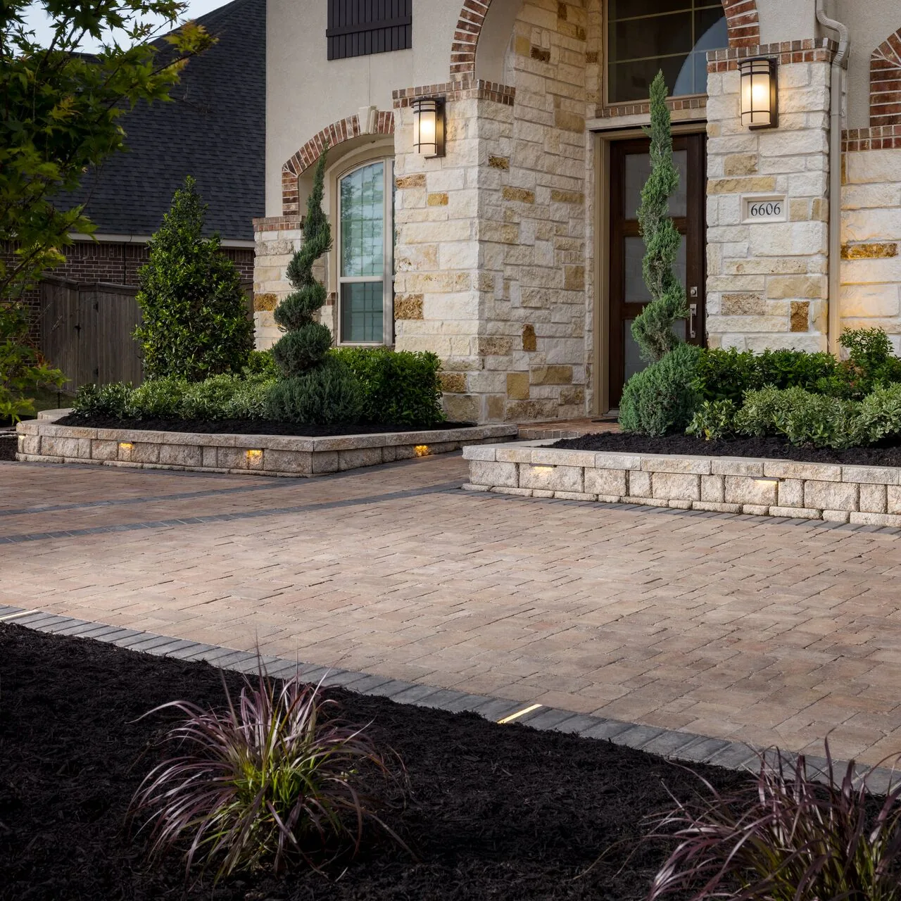 Warm-toned paved forecourt beside a stone house entrance and planted beds.