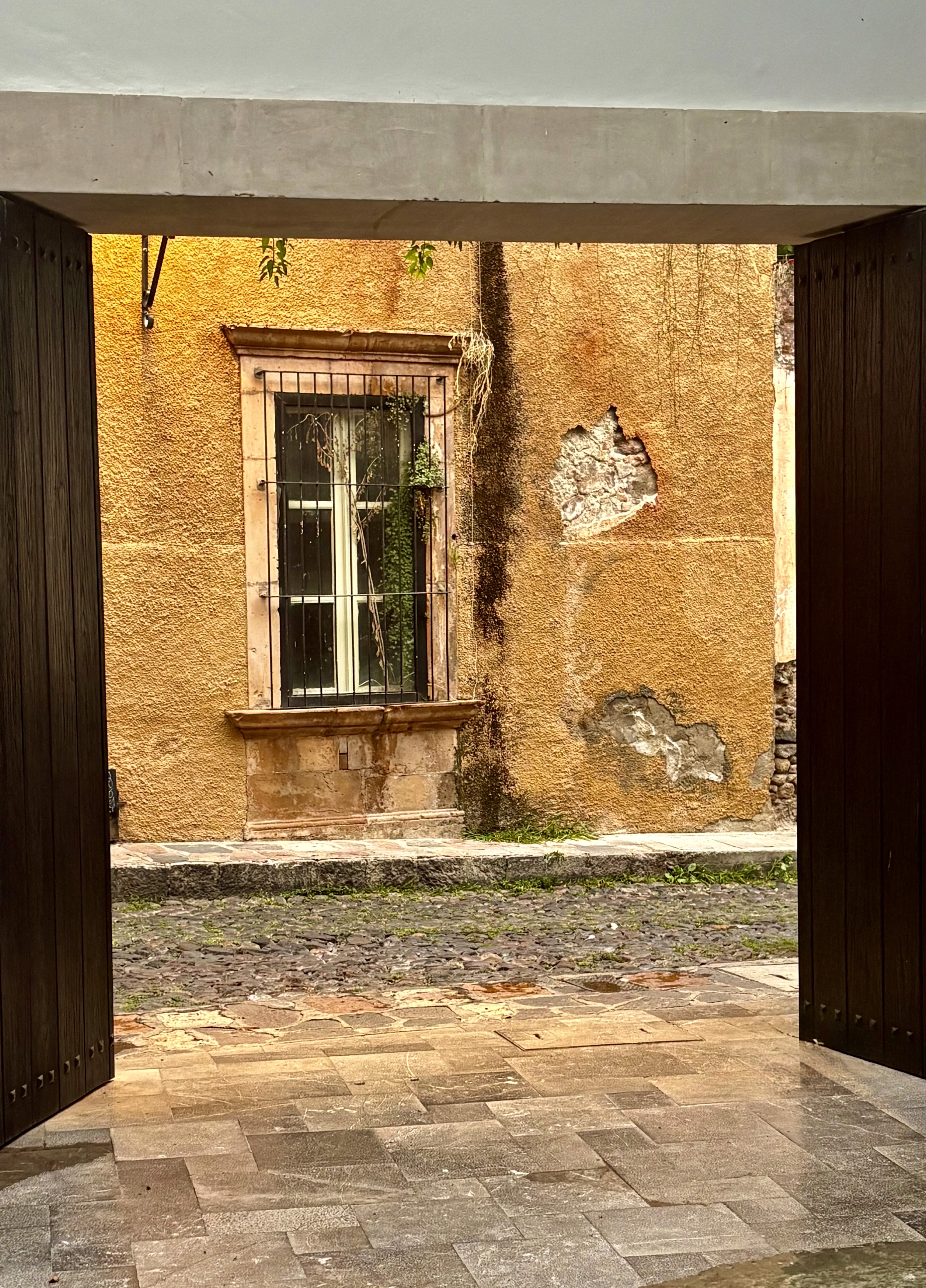 Weathered ochre wall and barred window framed by dark wooden doors in San Miguel de Allende