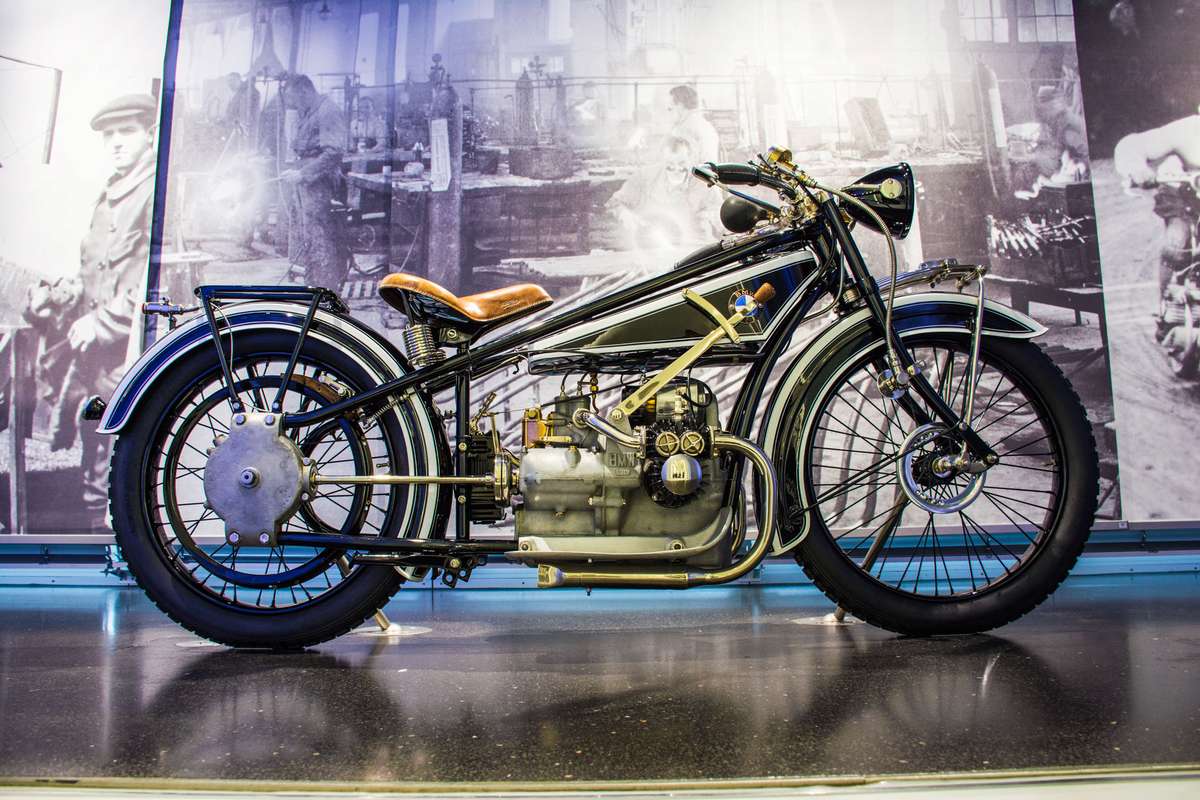 A customized cafe racer motorcycle with a black frame and tan seat sits in front of a large black-and-white mural.