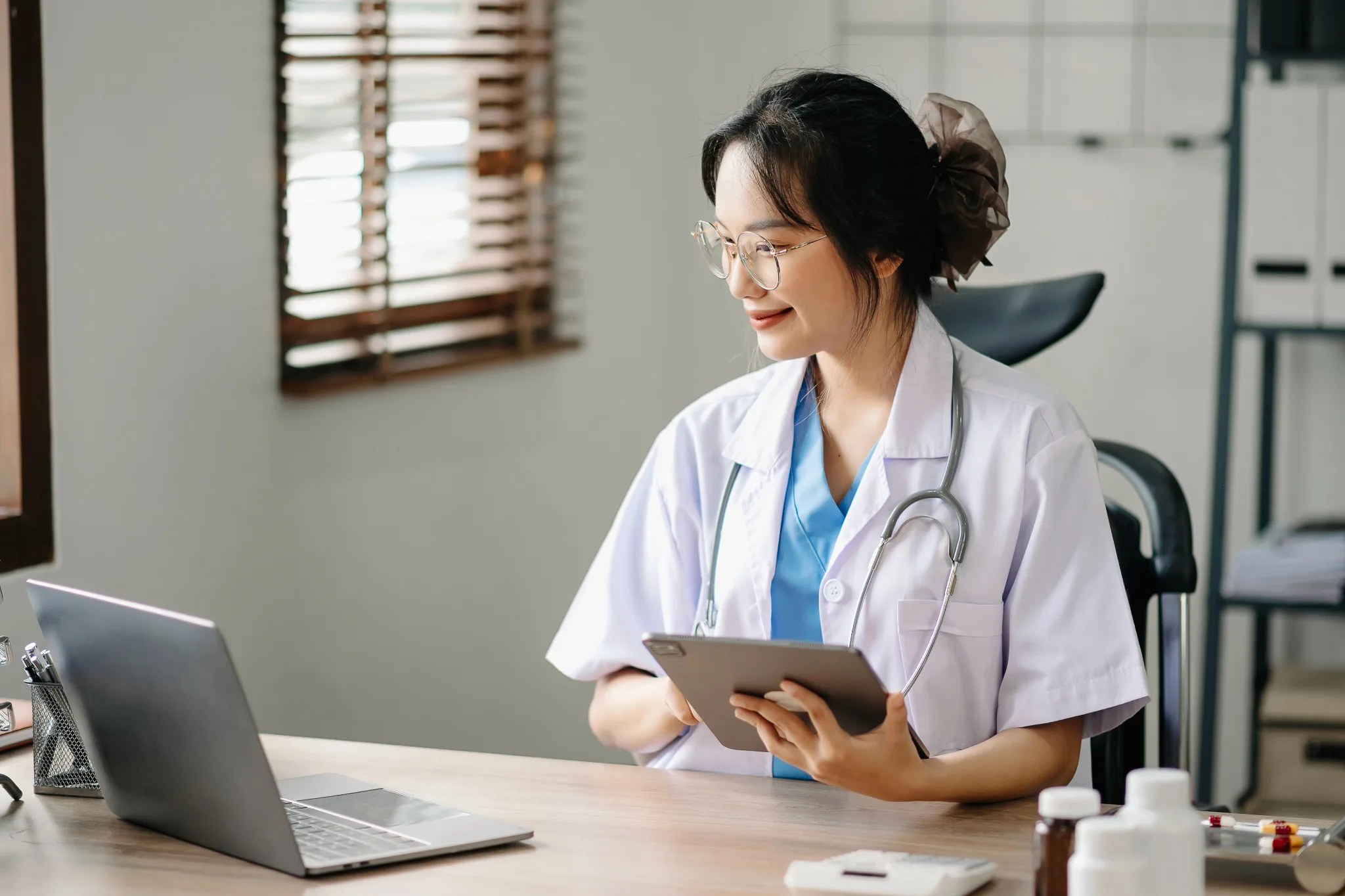 Smiling female physician, wearing a white coat with stethoscope around the neck, sitting on a chair with a table in front, holding a tablet and looking at a laptop.