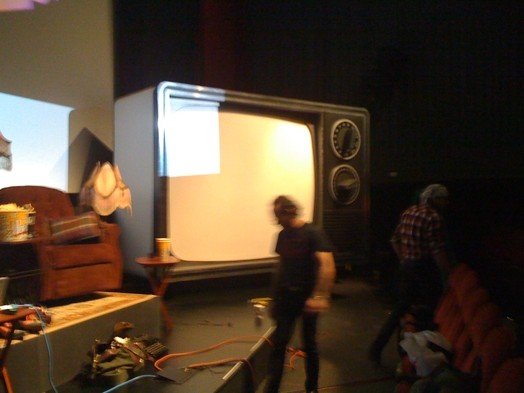 A photo of a big old TV set on a stage near an armchair and other show preps in a theatre.