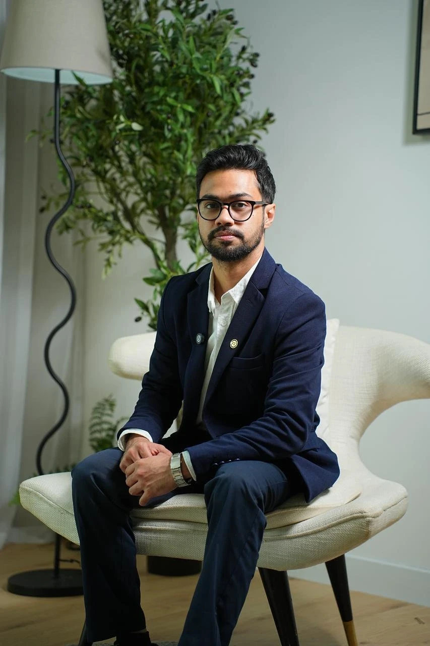 Manula Ranasinghe- Team Lead - Compliance - A man with glasses and a beard, wearing a navy blazer, sitting on a white chair, posing for a portrait.