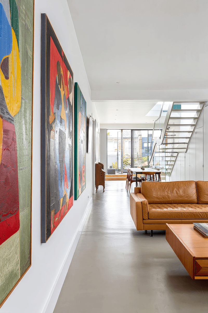 Modern minimalist hallway with colorful abstract art gallery wall, tan leather sofa, and floating staircase leading to natural light