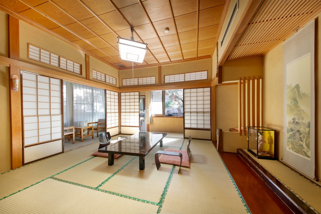 A bright, traditional Japanese room featuring tatami floors, a low central table with floor chairs, shoji screens, and a decorative alcove with a hanging scroll painting.