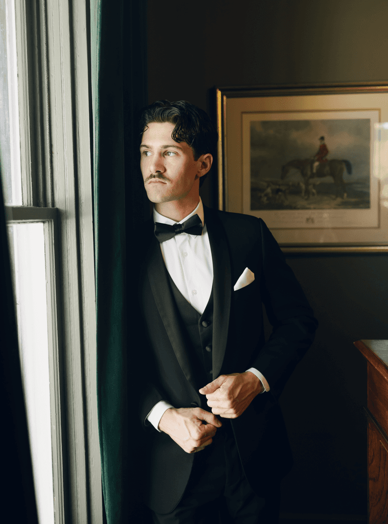     Palm Beach wedding photographer captures a groom adjusting his tuxedo beside a window in an editorial wedding portrait.