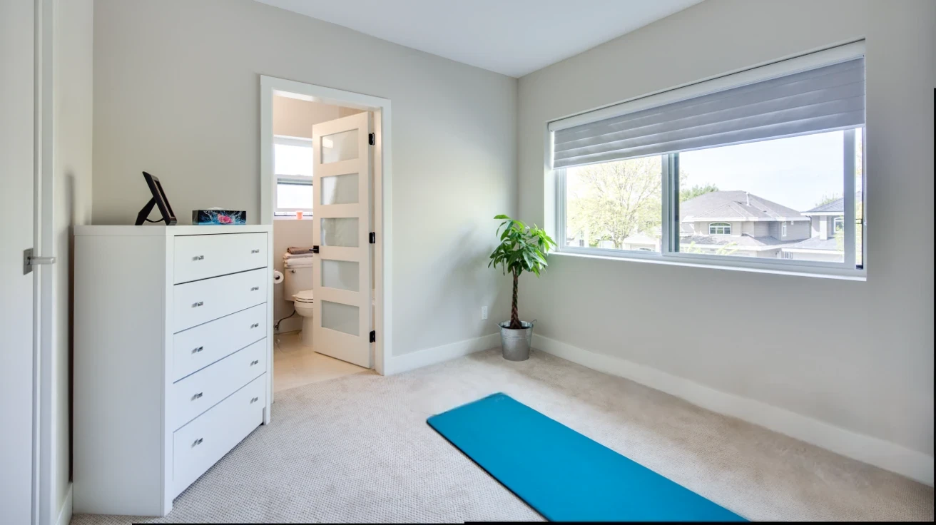 Bright bedroom with ensuite bathroom access, white dresser, exercise area with yoga mat, large window, and beige carpeting