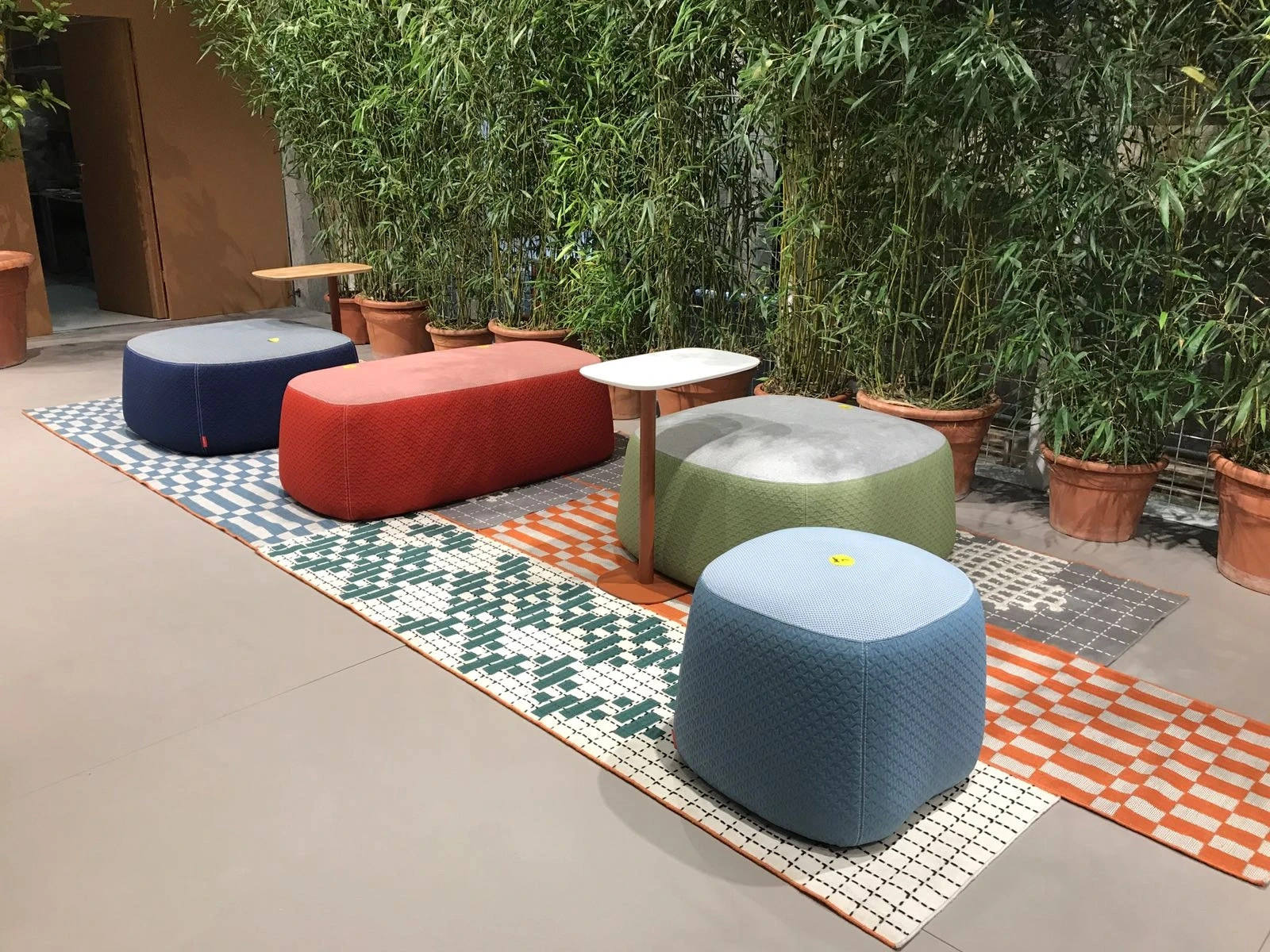 Quilted poufs in yellow, red, green and blue with small side tables on patterned runners in front of bamboo at the HAWORTH ORGATEC stand.