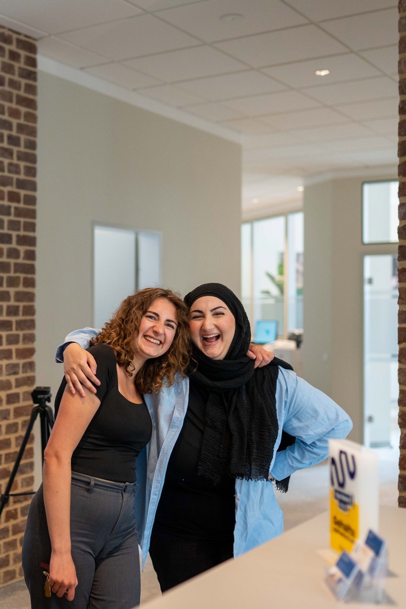 Zwei Mitarbeiterinnen von SLB stehen lachend nebeneinander in einem offenen Bürobereich und legen die Arme umeinander.